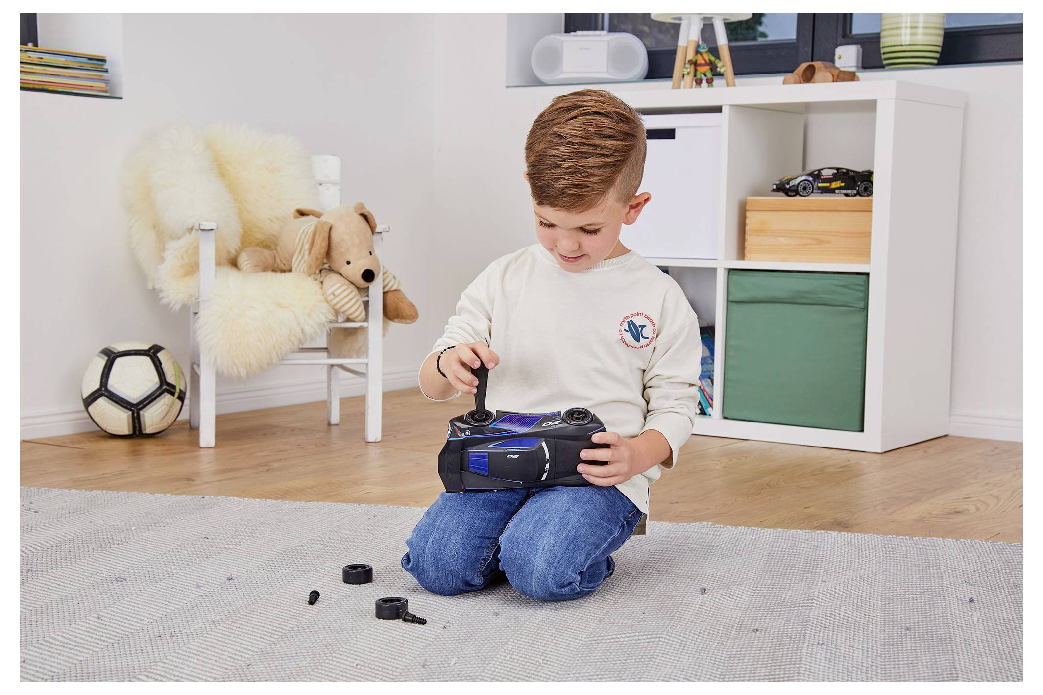Młody chłopiec klęczy na dywanie, składając samochód zdalnie sterowany wkrętakiem, otoczony zabawkami i pluszowym fotelem w pokoju zabaw.