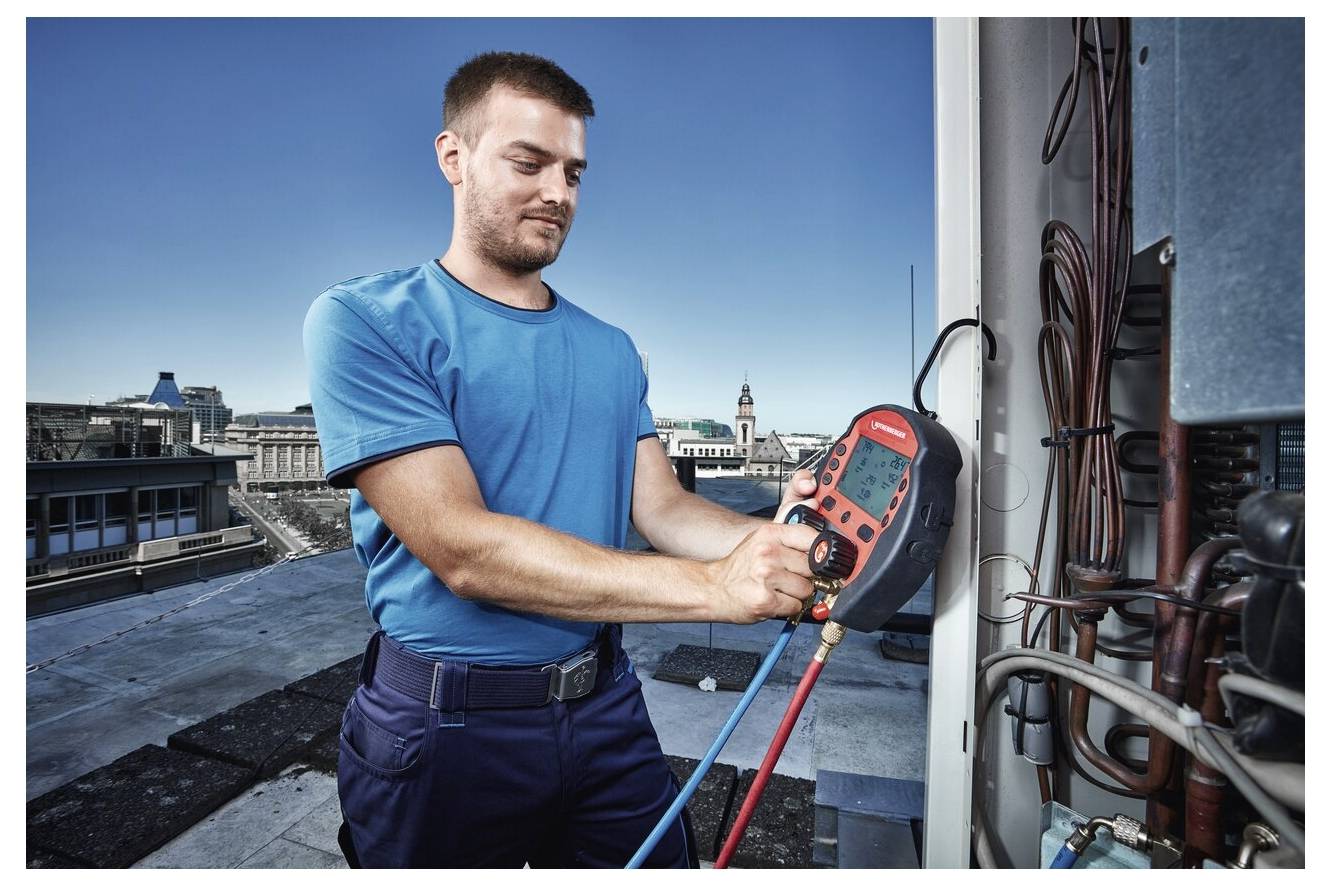 Technik używa cyfrowego manometru podczas konserwacji instalacji HVAC na dachu, otoczony budynkami miejskimi pod bezbłędnie czystym błękitnym niebem.