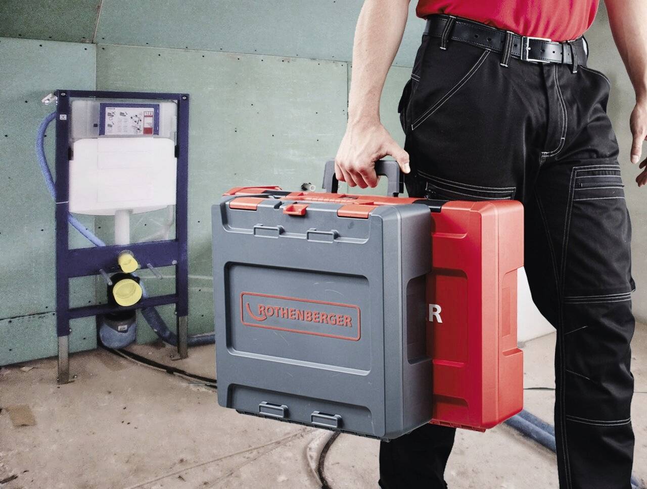 Osoba w stroju roboczym trzymająca czerwono-szare pudełko na narzędzia marki Rothenberger, stojąca w pobliżu sprzętu instalacyjnego w otoczeniu budowy.