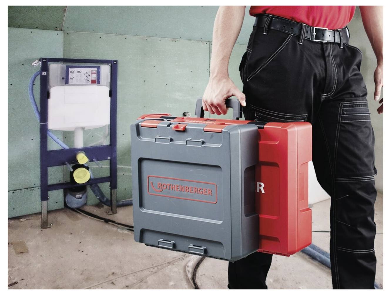 Osoba ubrana w strój roboczy niesie dwie skrzynki z narzędziami przed zamontowaną na ścianie instalacją hydrauliczną, co sugeruje naprawę lub montaż instalacji hydraulicznej.