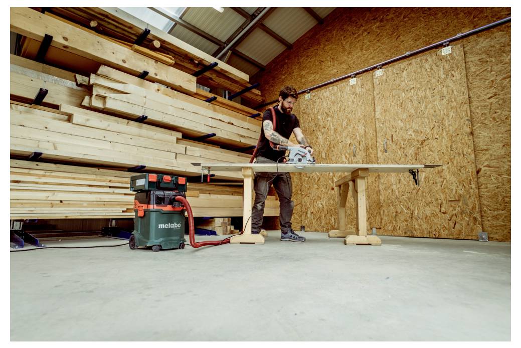 Osoba używa piły tarczowej do cięcia drewna na stole warsztatowym w warsztacie, w tle widać stosy desek.