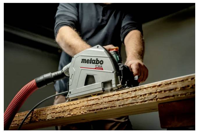 Osoba używająca piły tarczowej Metabo do cięcia drewnianej deski z podłączonym wężem do odkurzacza.
