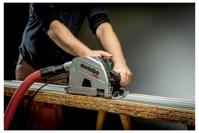 Osoba używająca piłę tarczową Metabo z wężem do odkurzacza do cięcia drewnianej deski na warsztacie pracy.