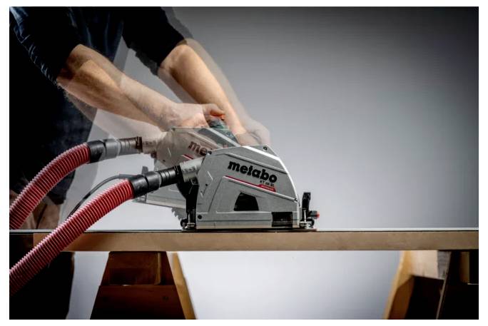 Osoba używająca narzędzia elektrycznego Metabo z czerwonymi wężami odkurzającymi do cięcia lub szlifowania wzdłuż krawędzi drewnianej deski na warsztacie.