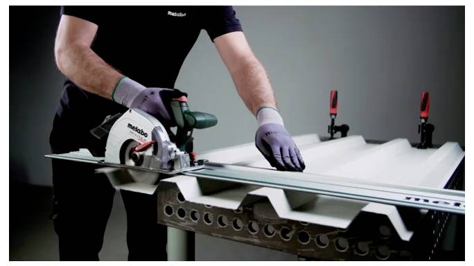 Osoba używająca piły tarczowej do cięcia arkusza metalu na stole roboczym, nosząca rękawice i trzymająca go za pomocą imadeł.