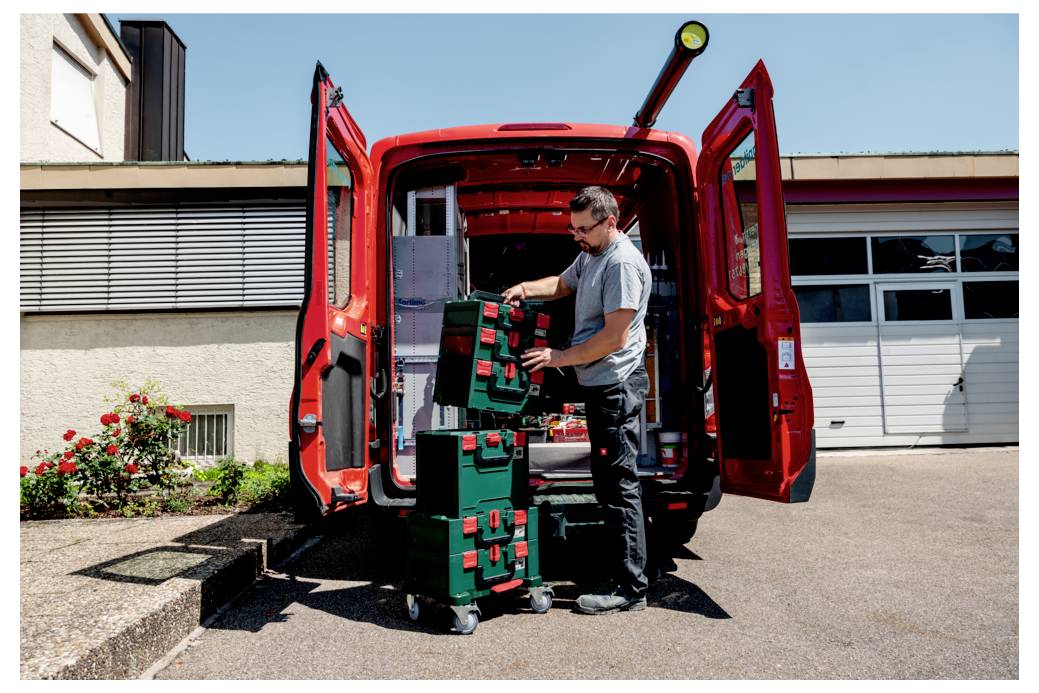 Osoba ładuje skrzynki narzędziowe do tylnej części czerwonego vana zaparkowanego przed budynkiem w słoneczny dzień, sugerując zadanie związane z pracą lub przeprowadzką.