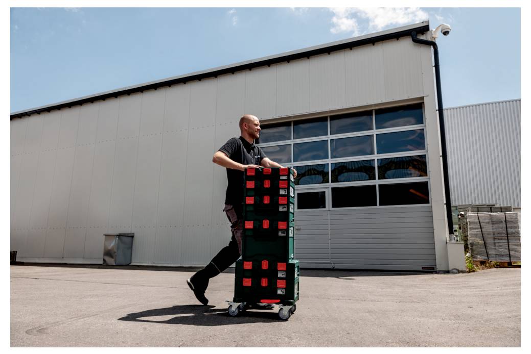 Osoba przesuwa stos zielonych skrzyń na wózku przed dużym budynkiem przemysłowym w słoneczny dzień.