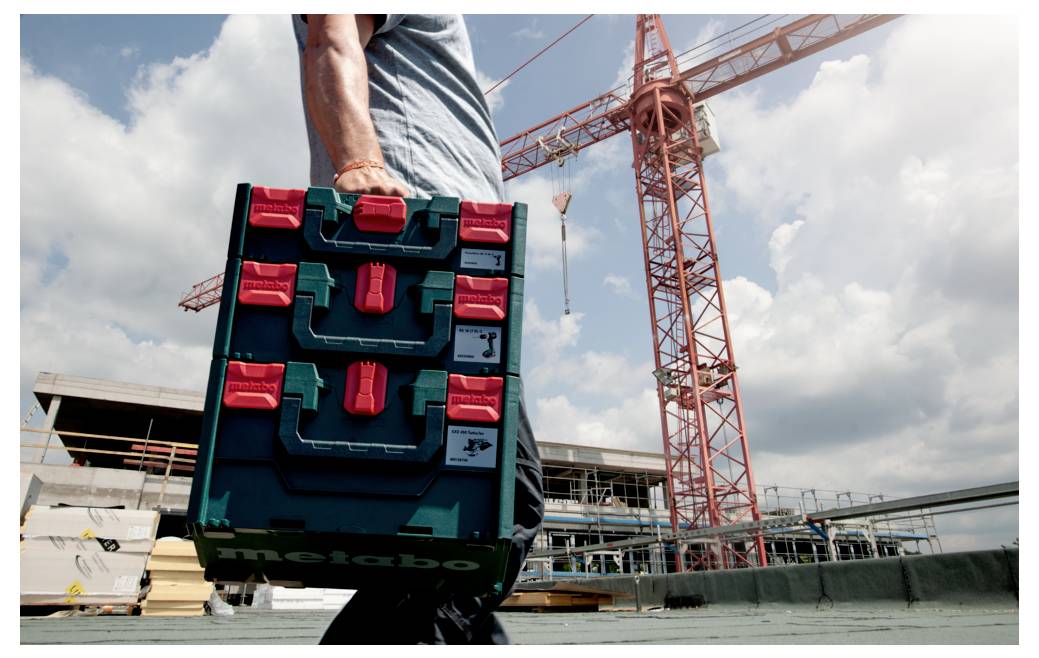 Osoba niesie piętrową skrzynkę narzędziową na placu budowy, z czerwonym dźwigiem i materiałami budowlanymi w tle, pod pochmurnym niebem.