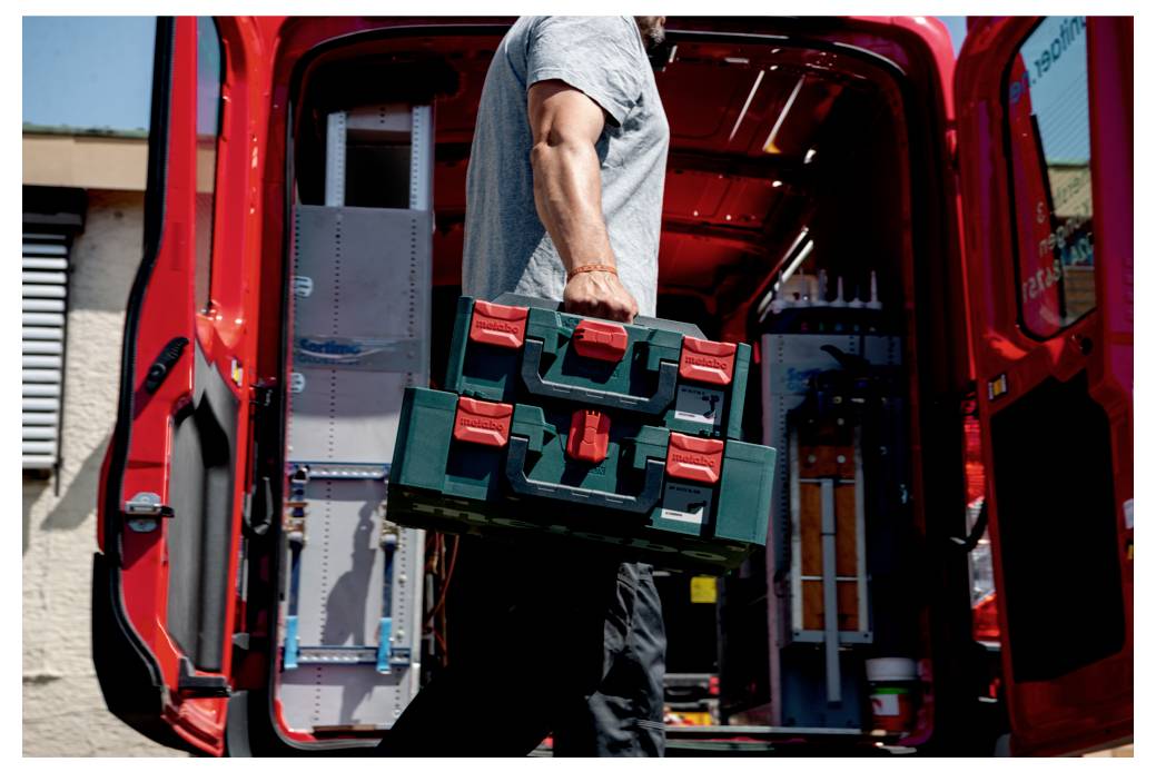 Osoba niosąca zielone pudełko z narzędziami zbliża się do otwartego czerwonego vana, prawdopodobnie rzemieślnik przygotowujący sprzęt do pracy.