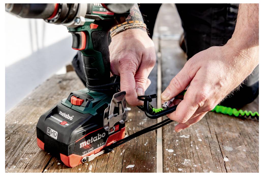 Osoba używa zielonej i czarnej wiertarki z zabezpieczonymi pasami bezpieczeństwa, co sugeruje koncentrację na bezpiecznych praktykach budowlanych.