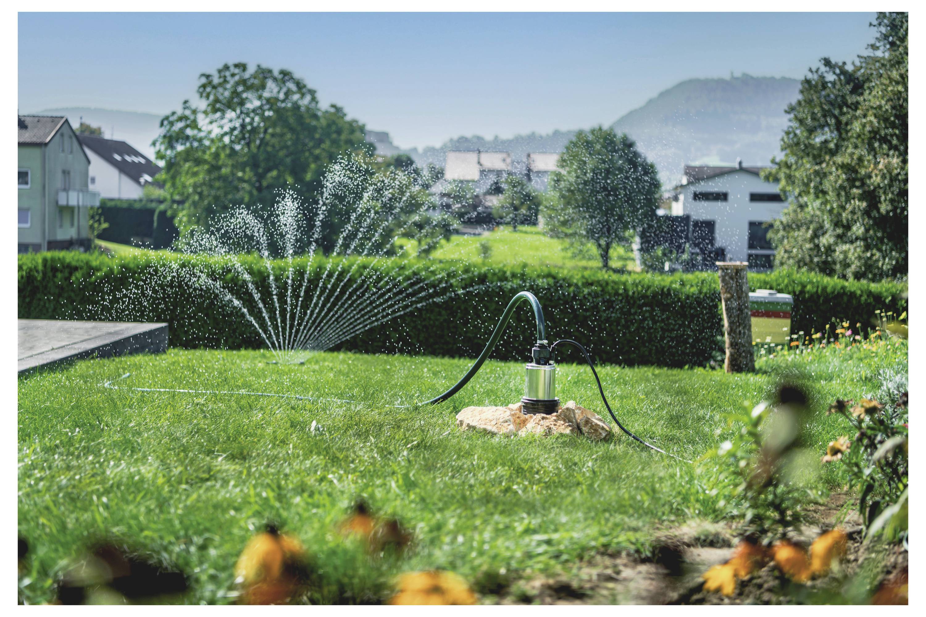 Zraszacz trawnikowy nawadnia ogród w dzielnicy mieszkaniowej, na tle domów i wzgórz, otoczony drzewami i kolorowymi kwiatami.