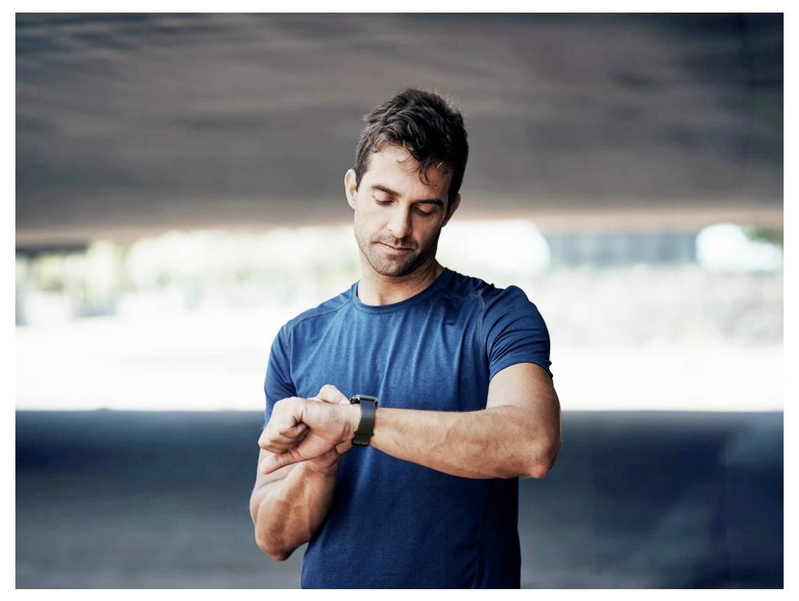 Mężczyzna w niebieskiej koszuli sprawdza zegarek na dworze, co sugeruje, że może śledzić czas lub monitorować swoją aktywność.