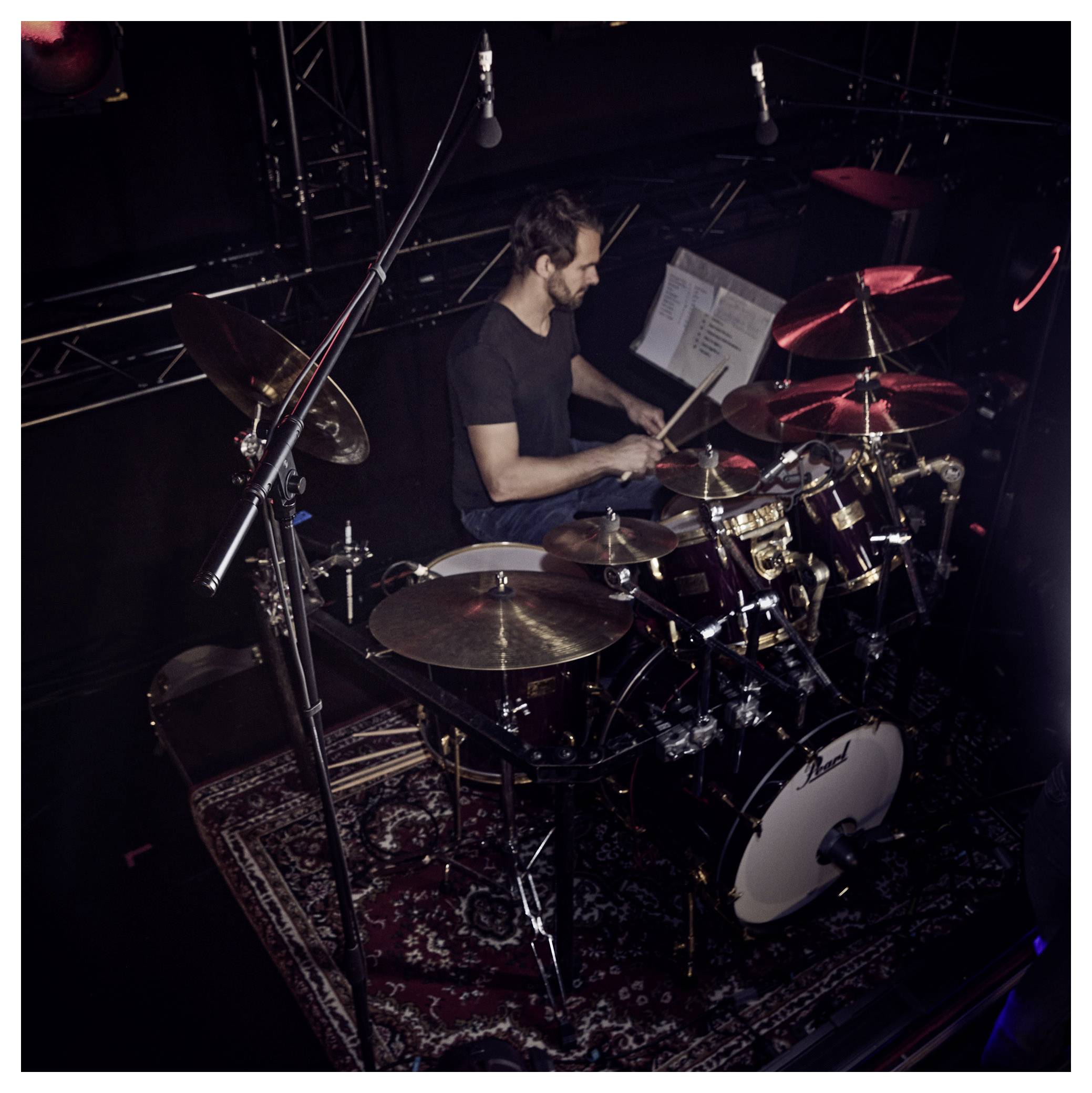 Mężczyzna gra na zestawie perkusyjnym na scenie, otoczony różnymi cymbałkami i mikrofonem, z widocznymi nutami na pulpicie.
