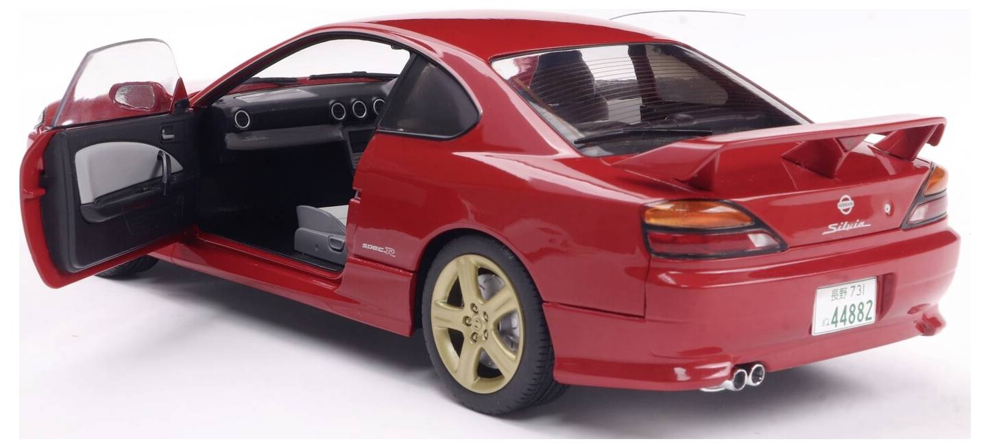 Czerwony samochód sportowy z otwartymi drzwiami kierowcy, złotymi felgami i tylnym skrzydłem, widziany od tyłu i z boku. Widoczna jest tablica rejestracyjna pojazdu.