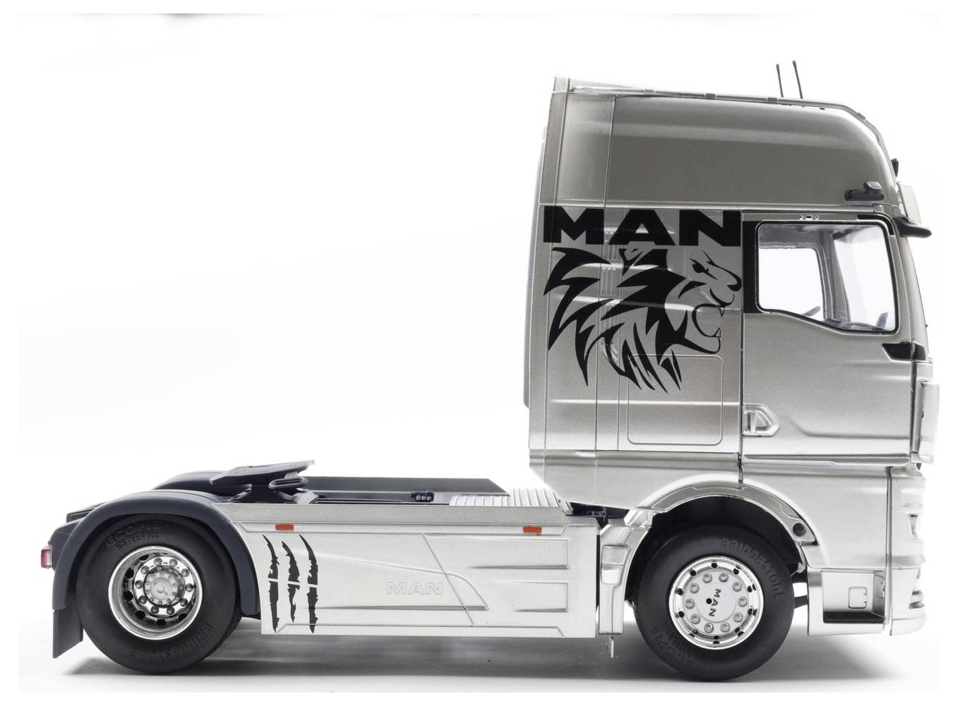Widoczna jest z boku srebrna kabina ciężarówki, na której znajduje się stylizowana grafika lwa oraz napis 'MAN' po stronie pojazdu. Ciężarówka nie ma widocznej przyczepy.