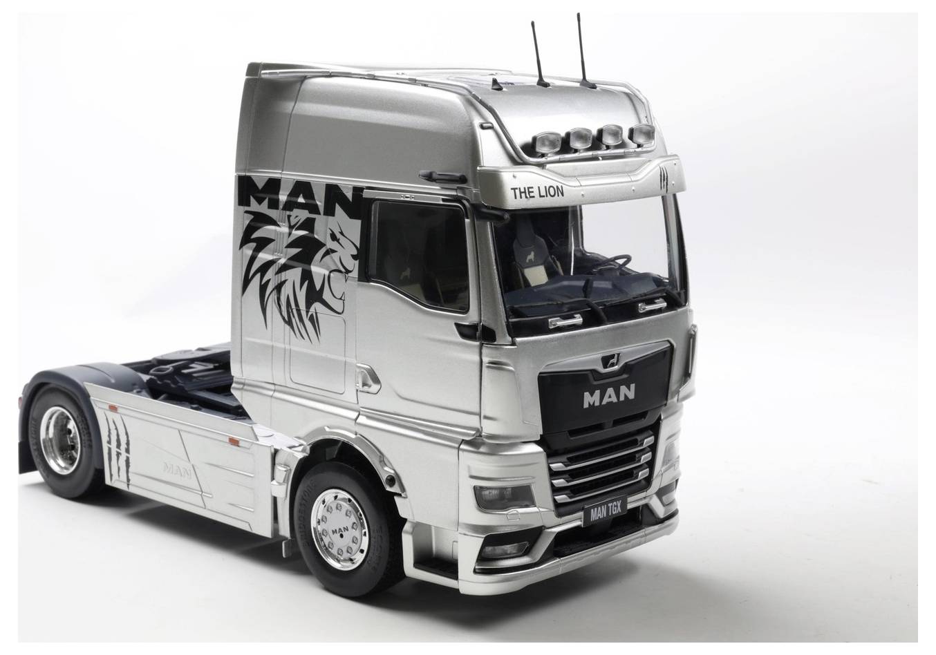 Srebrna ciężarówka z dokładnie namalowaną grafiką lwa na boku, oznaczona napisem 'MAN - LWIE OBLICZE', stoi na białym, jednolitym tle.