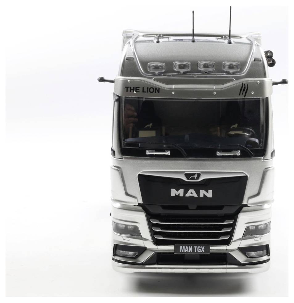 Srebrny samochód ciężarowy MAN TGX jest pokazany od przodu z napisem "THE LION" na górze. Ciężarówka posiada wiele świateł na dachu.