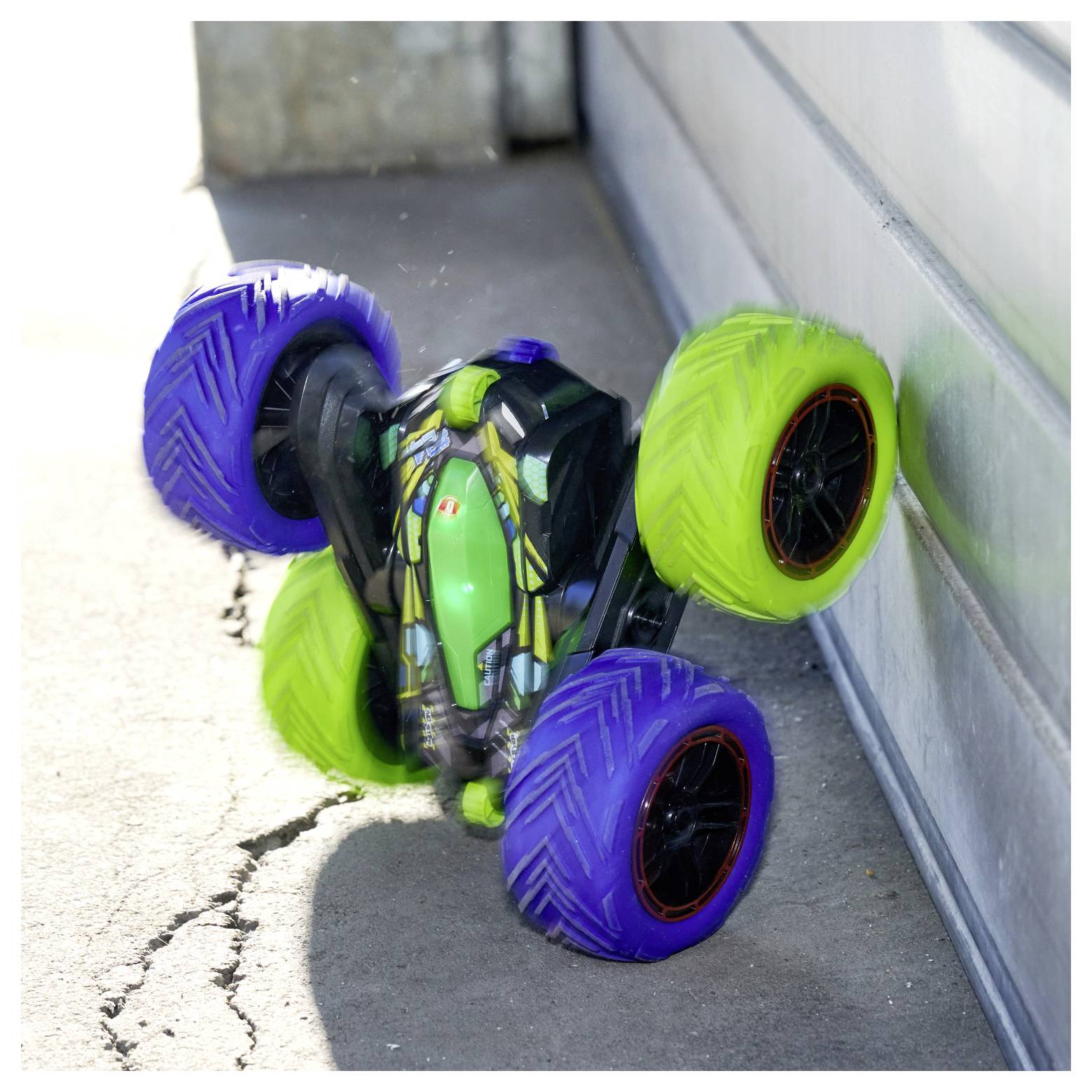 Zdalnie sterowany samochód przewraca się na bok, ukazując żywe, fioletowe i zielone koła, na betonowej powierzchni na wolnym powietrzu, w pobliżu budynku.