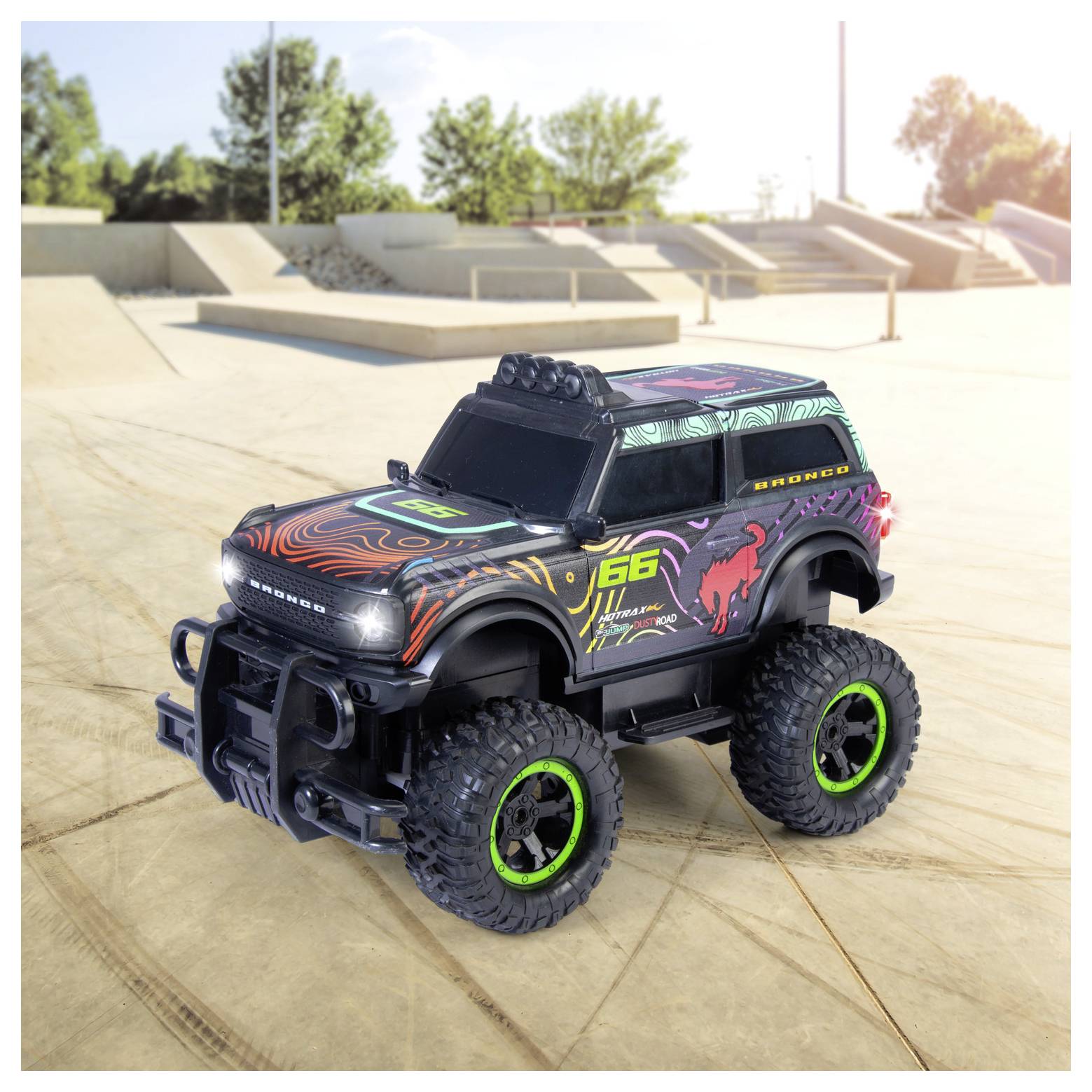 Kolorowy zabawkowy pojazd terenowy z dużymi oponami i przygodowymi naklejkami na betonowej powierzchni, z drzewami i rampami w tle.