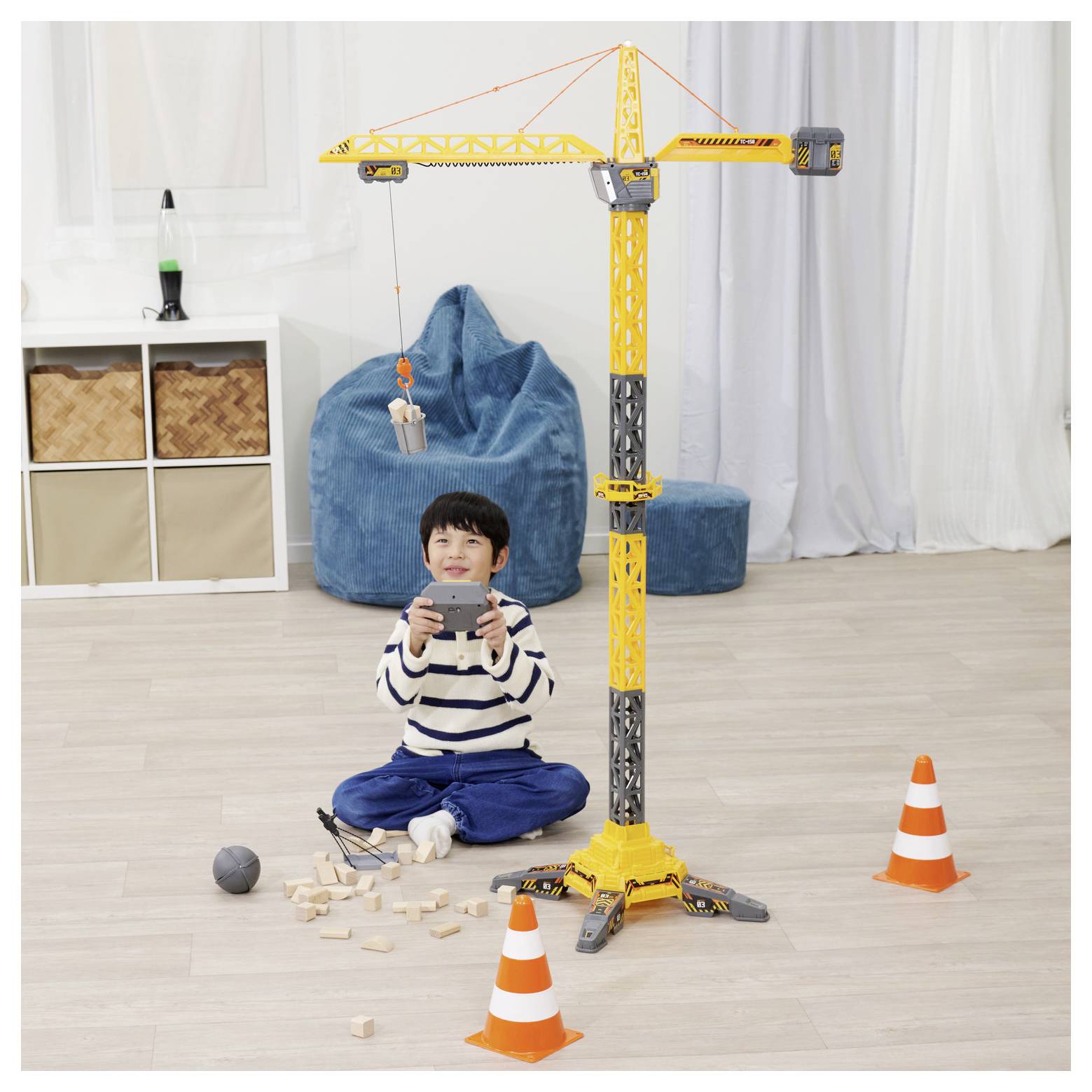 Dziecko bawi się zabawkowym dźwigiem budowlanym w pomieszczeniu, otoczone drewnianymi klockami i pomarańczowymi pachołkami drogowymi.