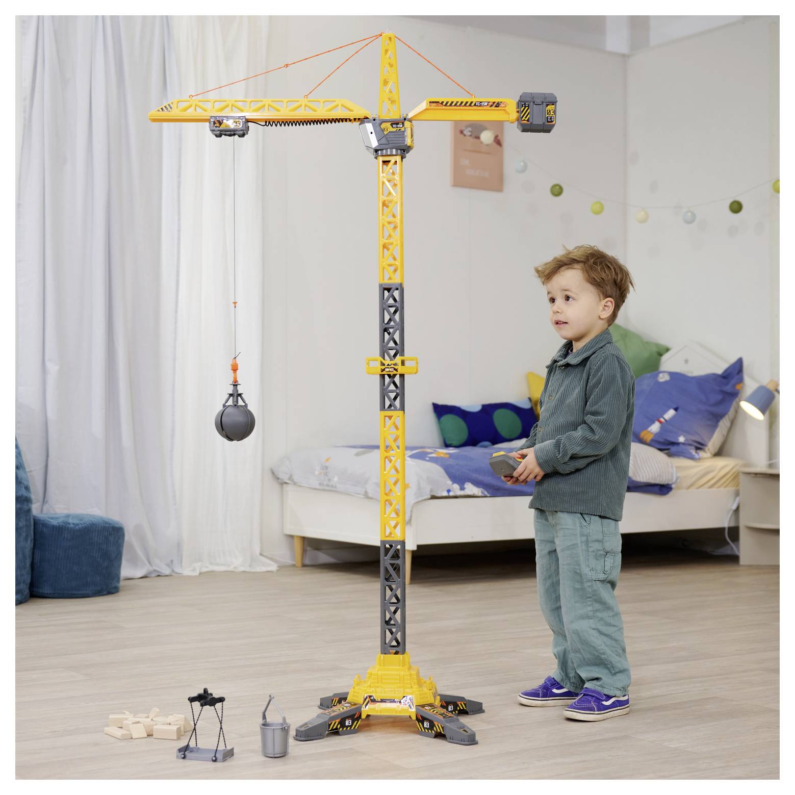 Młody chłopiec bawi się dużym zabawkowym dźwigiem budowlanym w sypialni. Dźwig posiada przystawkę z kulą wyburzeniową, a pokój jest urządzony do zabawy.
