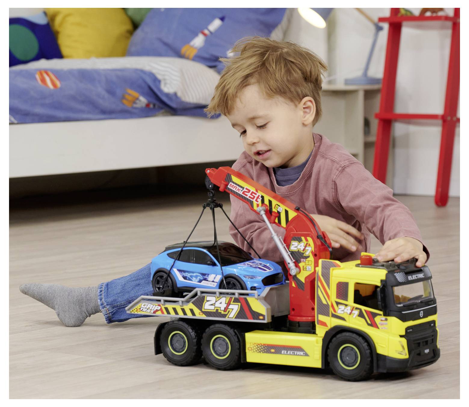 Dziecko bawi się zabawkowym holownikiem w sypialni, używając go do podniesienia niebieskiego samochodu zabawki leżącego na podłodze.