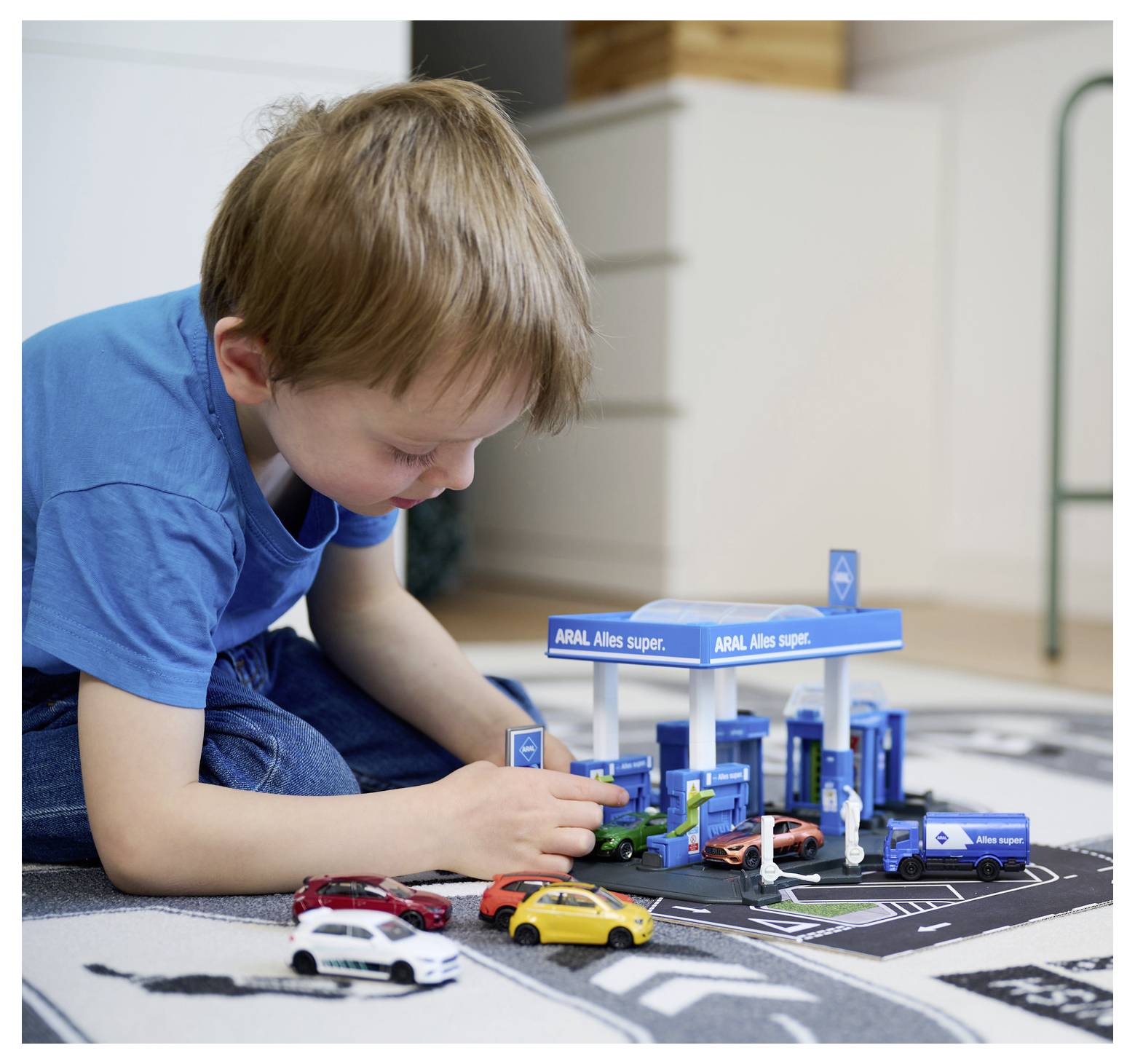 Dziecko bawi się zabawkowymi samochodami na miniaturowej stacji benzynowej. Stacja posiada markę 'ARAL' i kolorowe samochodziki. Scena jest pełna zabawy i wyobraźni.