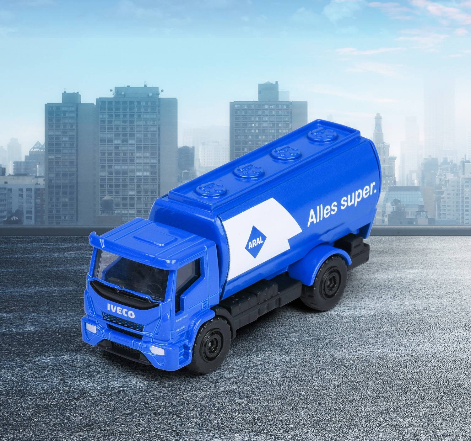Niebieska zabawkowa ciężarówka marki 'Iveco' z logo 'Aral' stoi na dachu z panoramą miasta w tle, a na niej widnieje napis 'Alles super.'