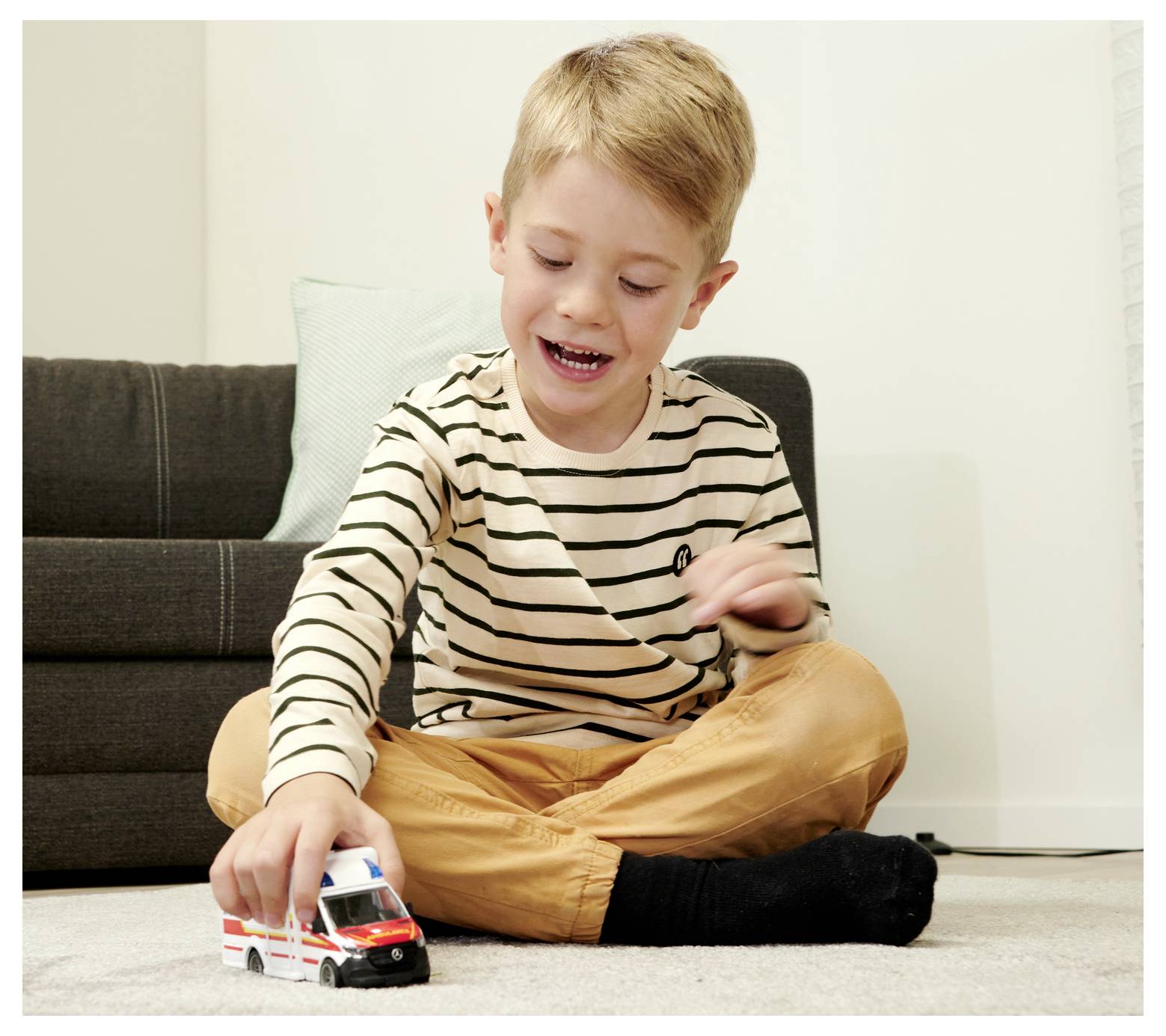 Mały chłopiec siedzący na dywanie bawi się radośnie zabawkową karetką pogotowia, wykazując się twórczą wyobraźnią.