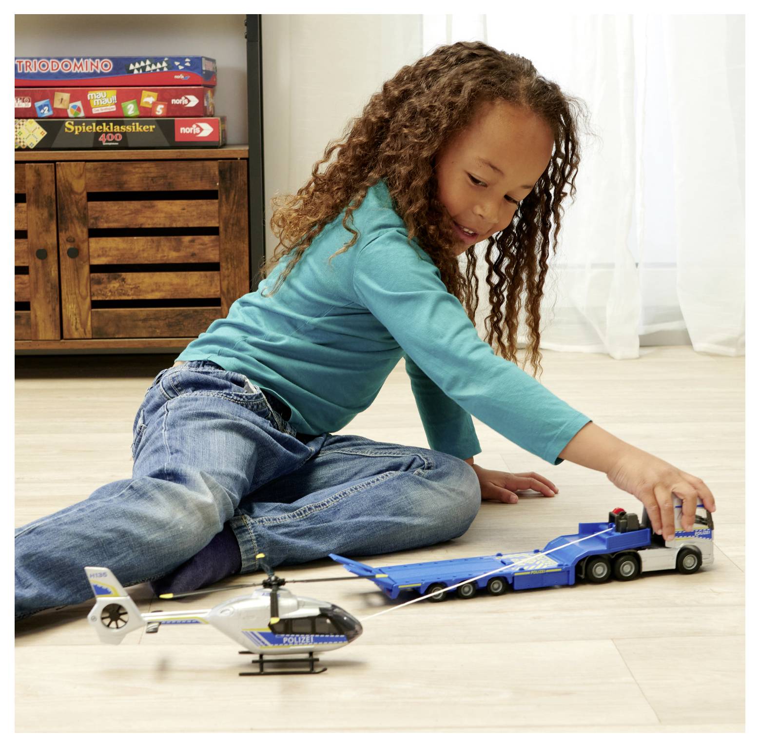 Dziecko w niebieskiej koszulce bawi się na podłodze zabawkowymi pojazdami, w tym zabawkowym helikopterem i ciężarówką, w pobliżu szafki z grami planszowymi.