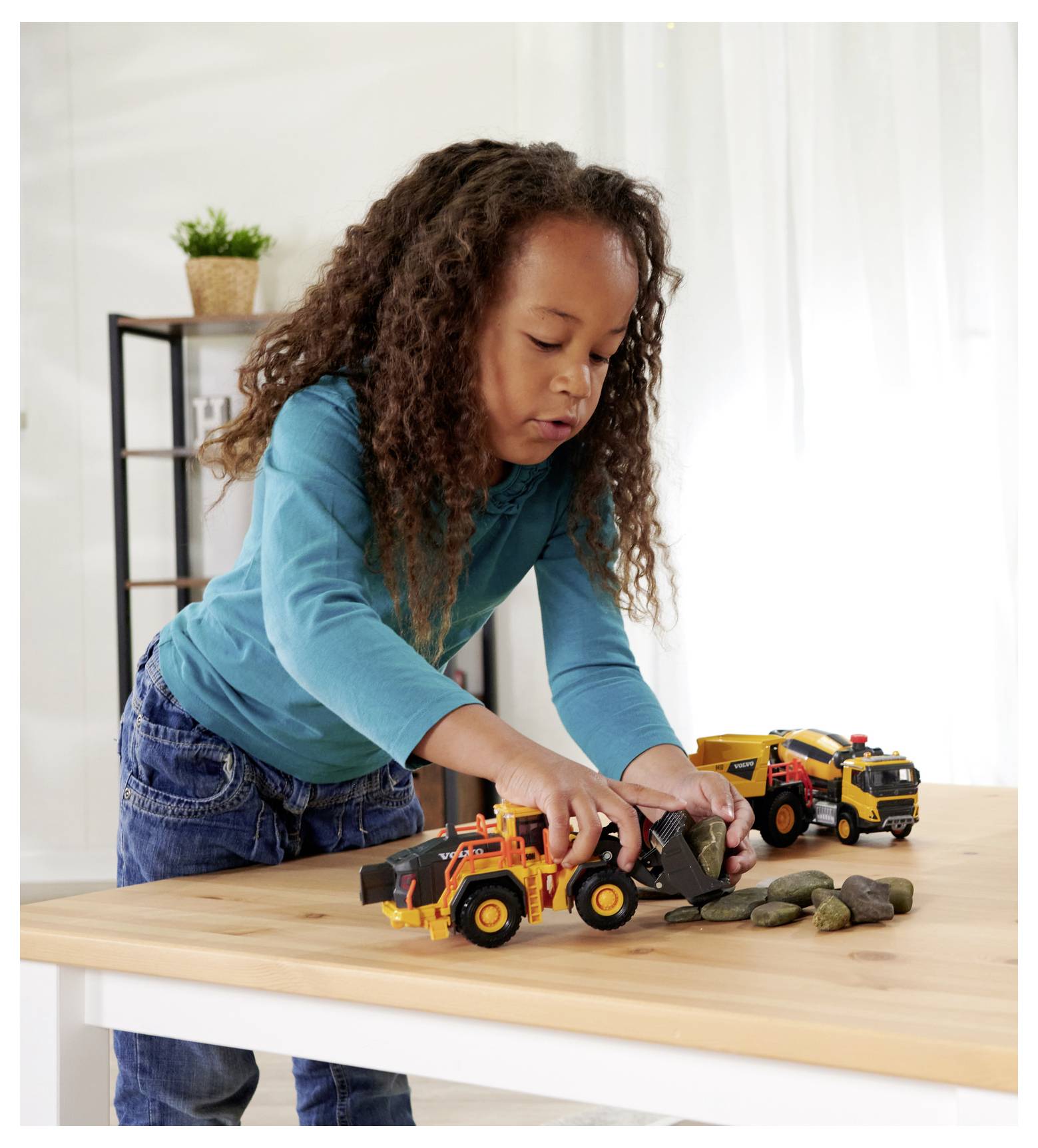Dziecko bawi się miniaturowymi pojazdami budowlanymi na drewnianym stole, układając małe kamyki. Scena rozgrywa się w jasnym pomieszczeniu z rośliną i regałem na książki.