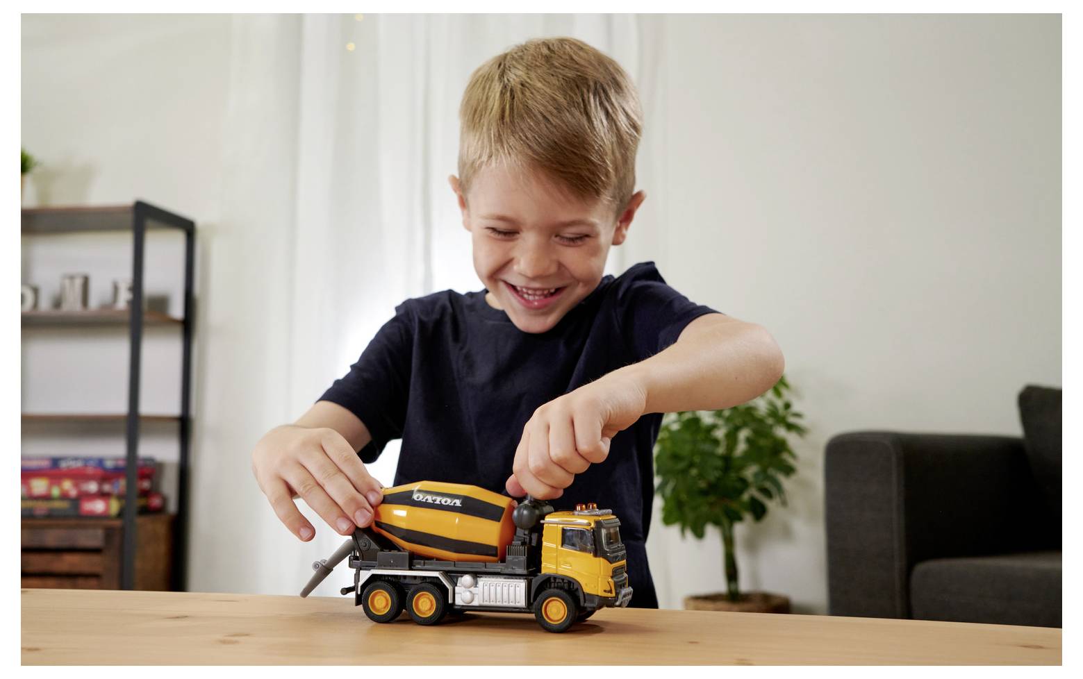 Dziecko o jasnobrązowych włosach bawi się zabawkową betoniarką na stole, uśmiechając się radośnie. W tle widać roślinę i regał na książki.