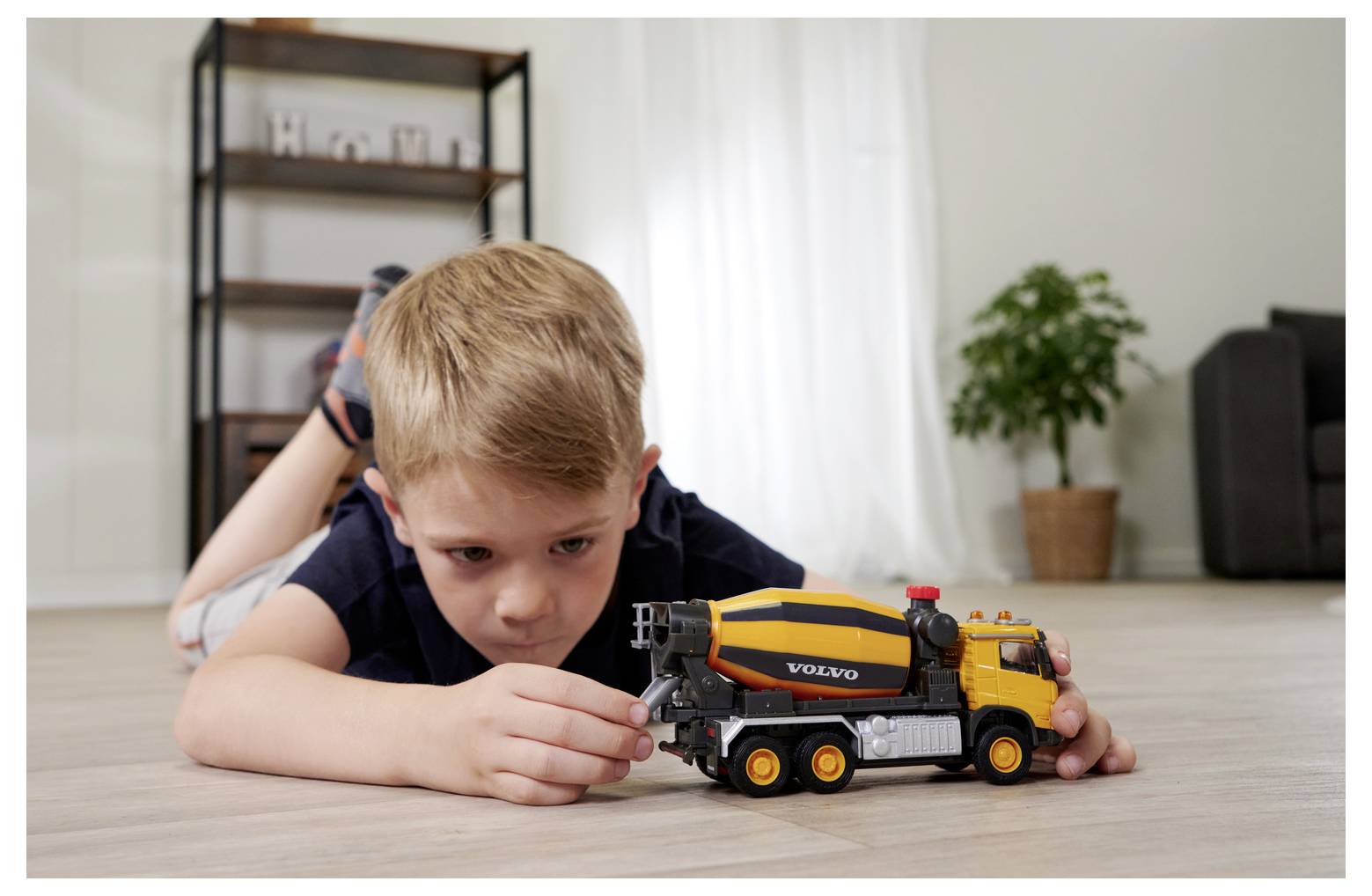 Młody chłopiec leży na podłodze, zaabsorbowany zabawą miniaturową zabawkową ciężarówką do mieszania cementu w salonie.