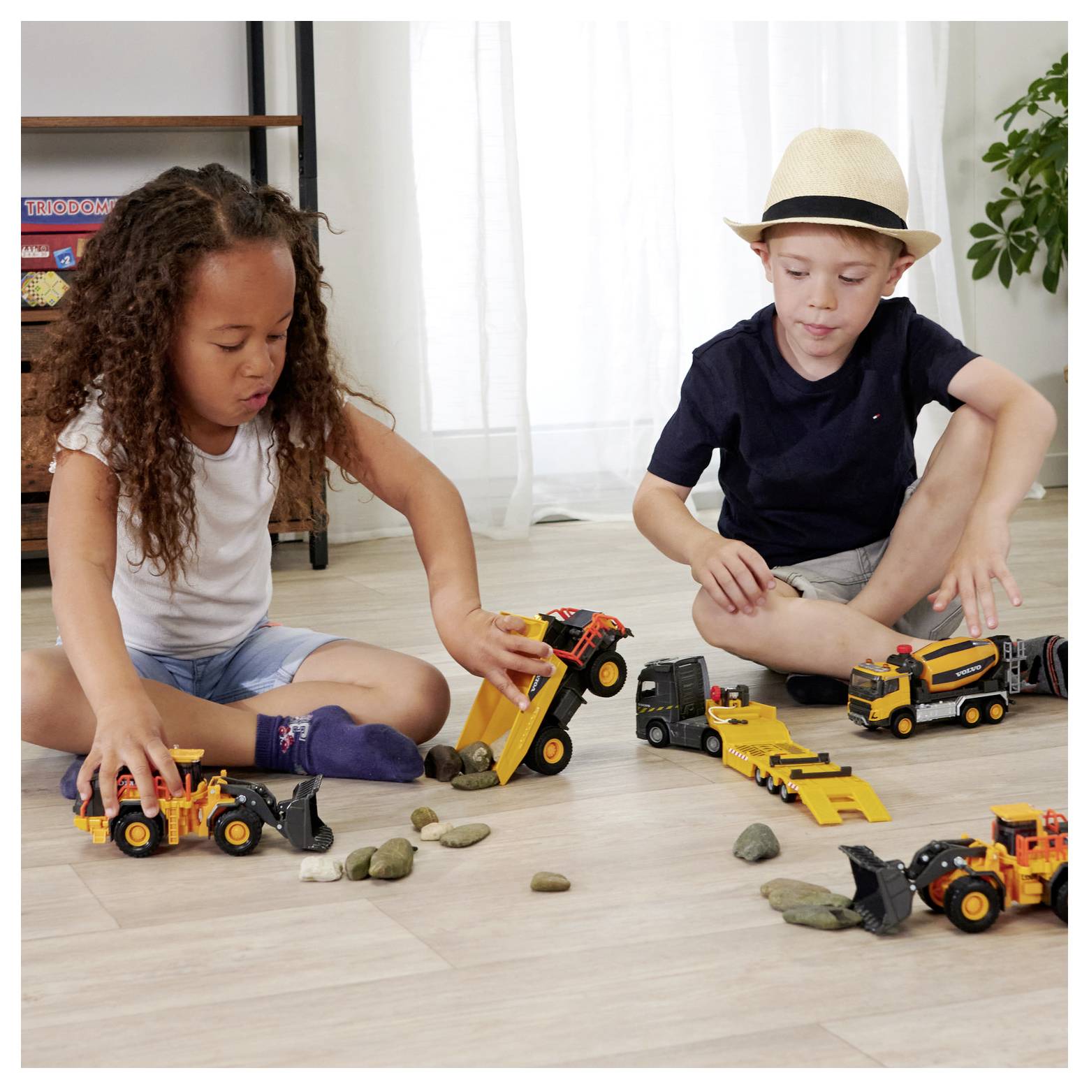 Dwoje dzieci siedzi na drewnianej podłodze, bawiąc się zabawkowymi pojazdami budowlanymi i kamieniami. Na tle widoczny jest doniczkowy kwiat oraz półka.