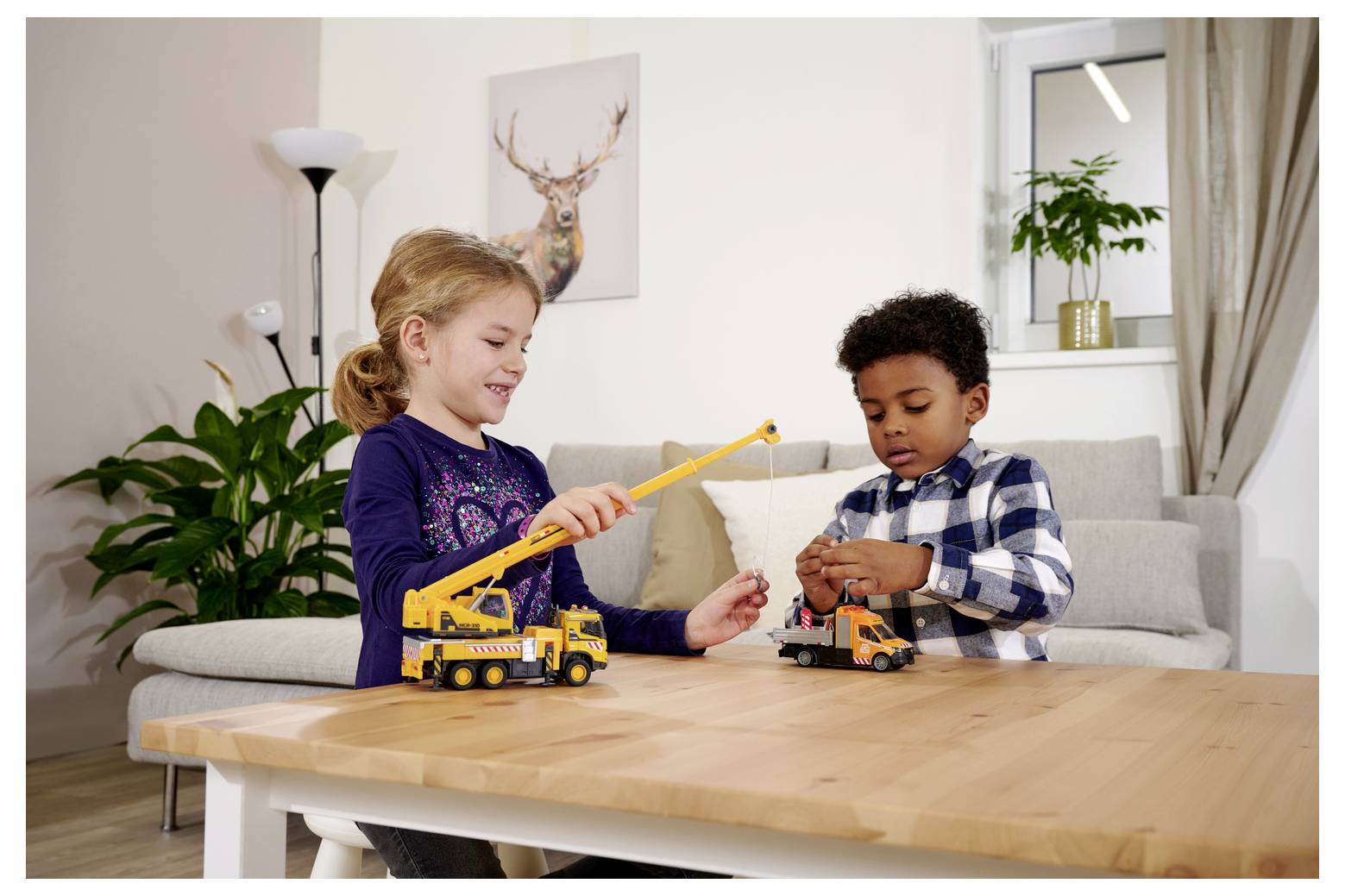 Dwoje dzieci bawi się zabawkowymi pojazdami budowlanymi przy drewnianym stole w salonie, z kanapą i rośliną w tle.