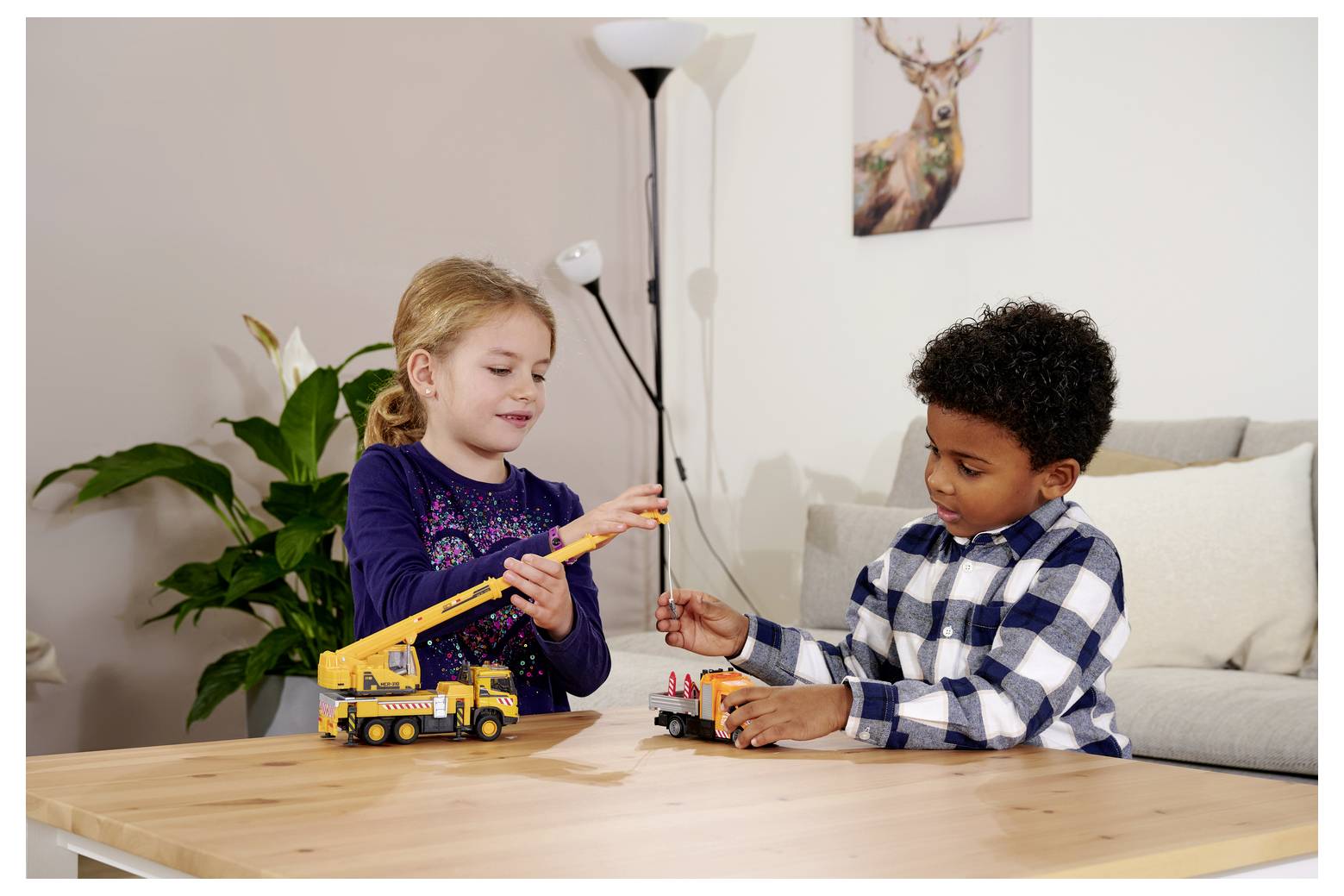 Dwoje dzieci bawi się zabawkowymi pojazdami budowlanymi na drewnianym stole w salonie. Na tle widać roślinę, lampę i obraz z jeleniem.
