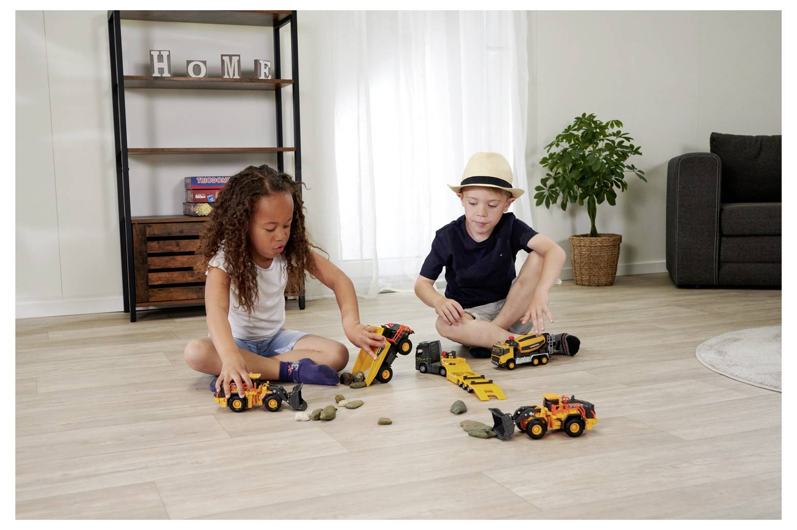 Dzieci bawią się w pomieszczeniu zabawkowymi pojazdami budowlanymi na drewnianej podłodze, otoczone małymi kamieniami. Na tle widać półkę z napisem „Dom".