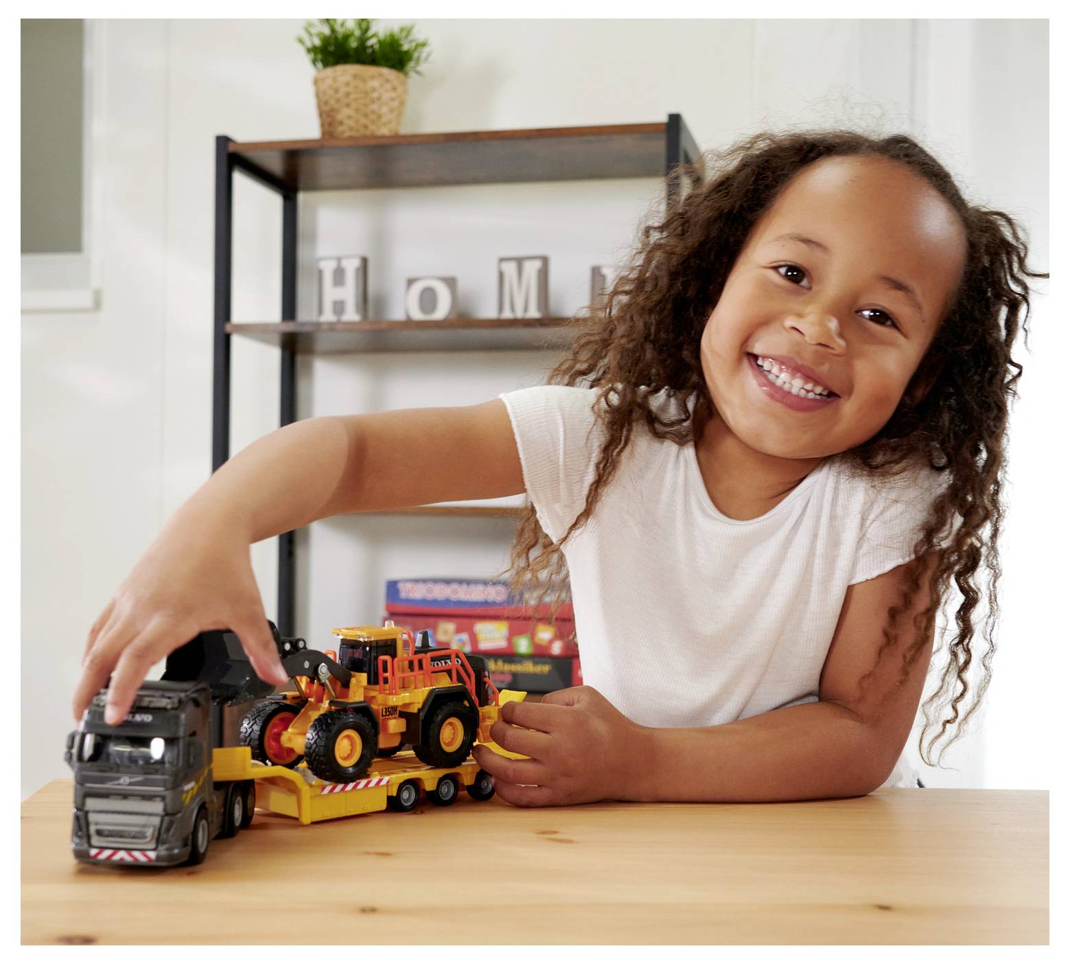 Uśmiechnięte dziecko bawi się zabawkowymi ciężarówkami na stole, a w tle widać półkę i elementy wystroju wnętrza.