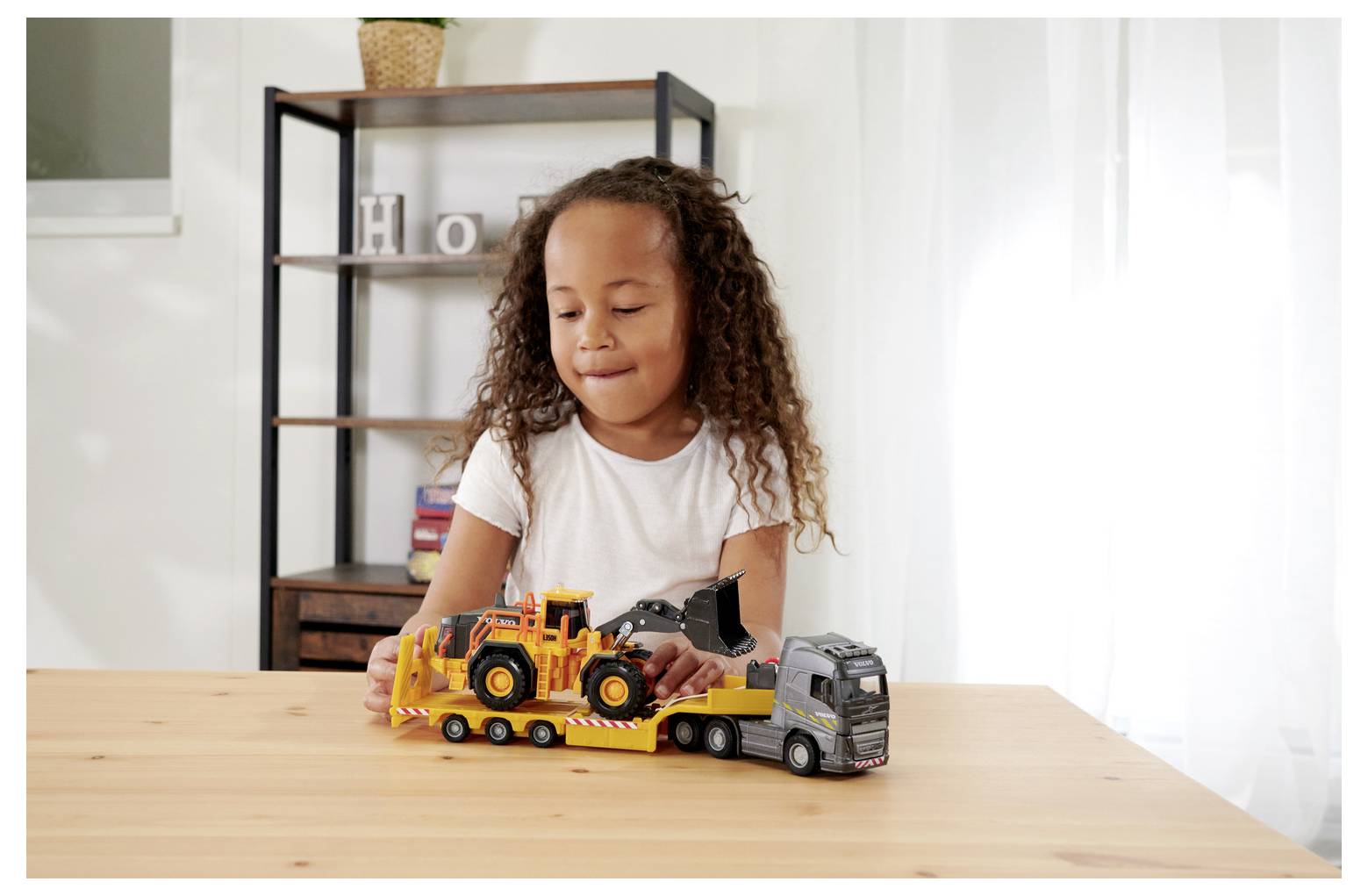 Dziecko bawi się zabawkowymi pojazdami budowlanymi na drewnianym stole w jasno oświetlonym pokoju, na tle którego widać półkę.