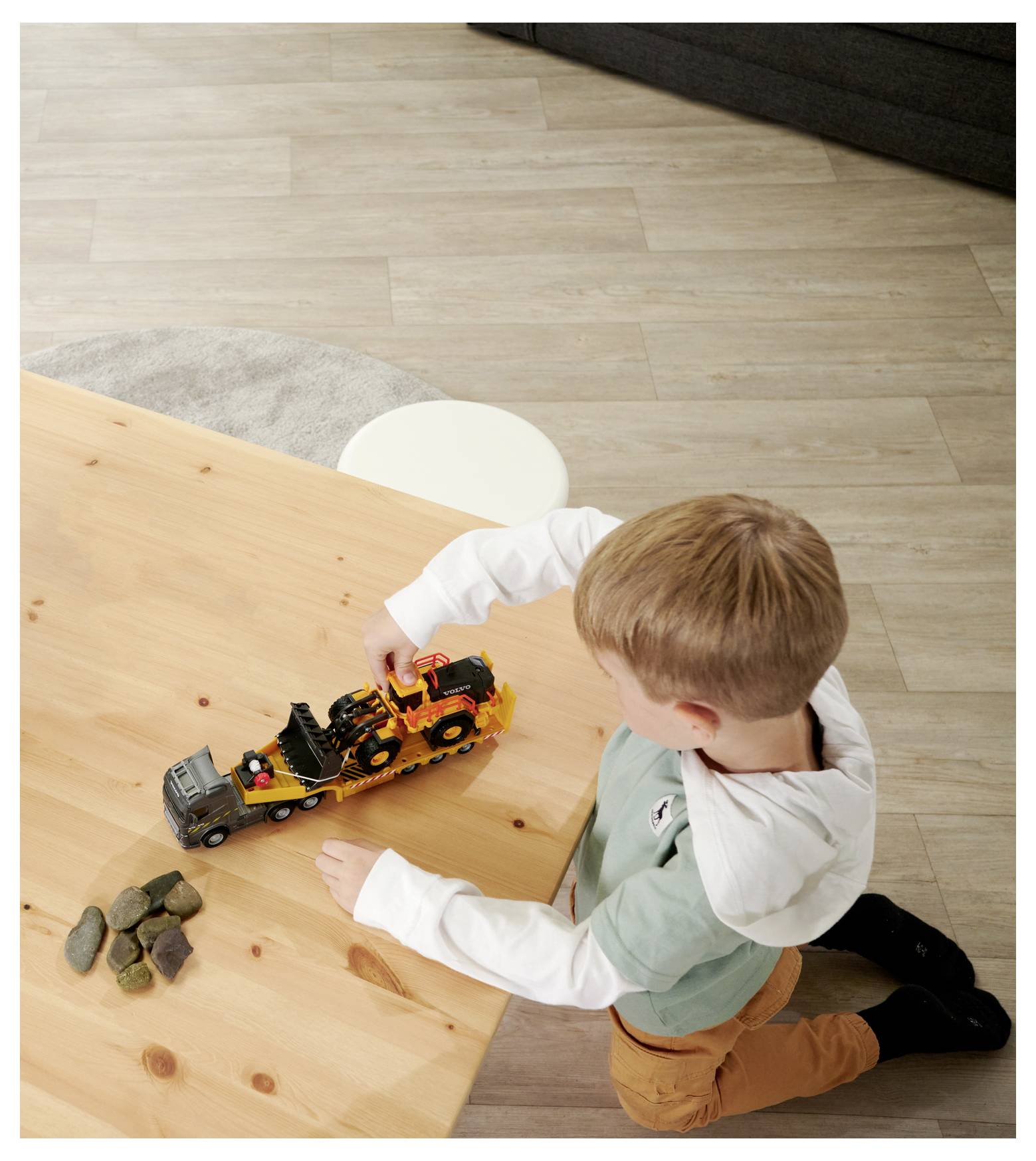 Dziecko siedzi na podłodze, bawiąc się zabawkowymi pojazdami budowlanymi na drewnianym stole, w sąsiedztwie małej sterty kamieni.