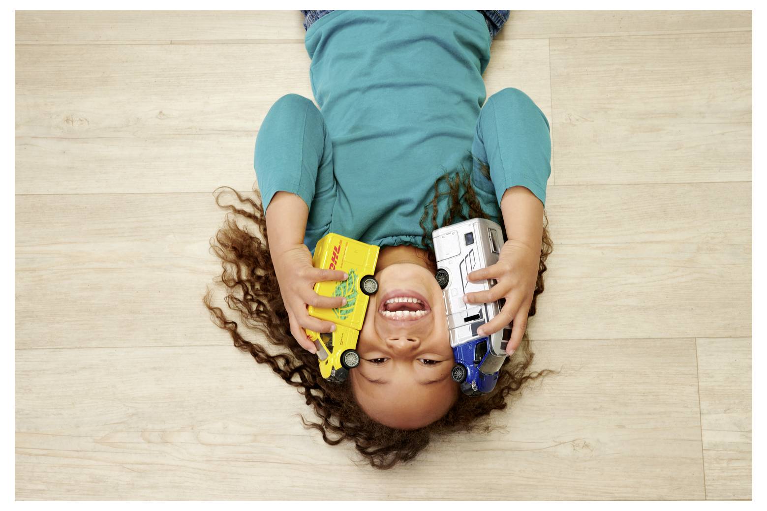 Dziecko leżące na drewnianej podłodze, uśmiechnięte, przytulające do policzków zabawkowe autobusy, jeden żółty, jeden biały.