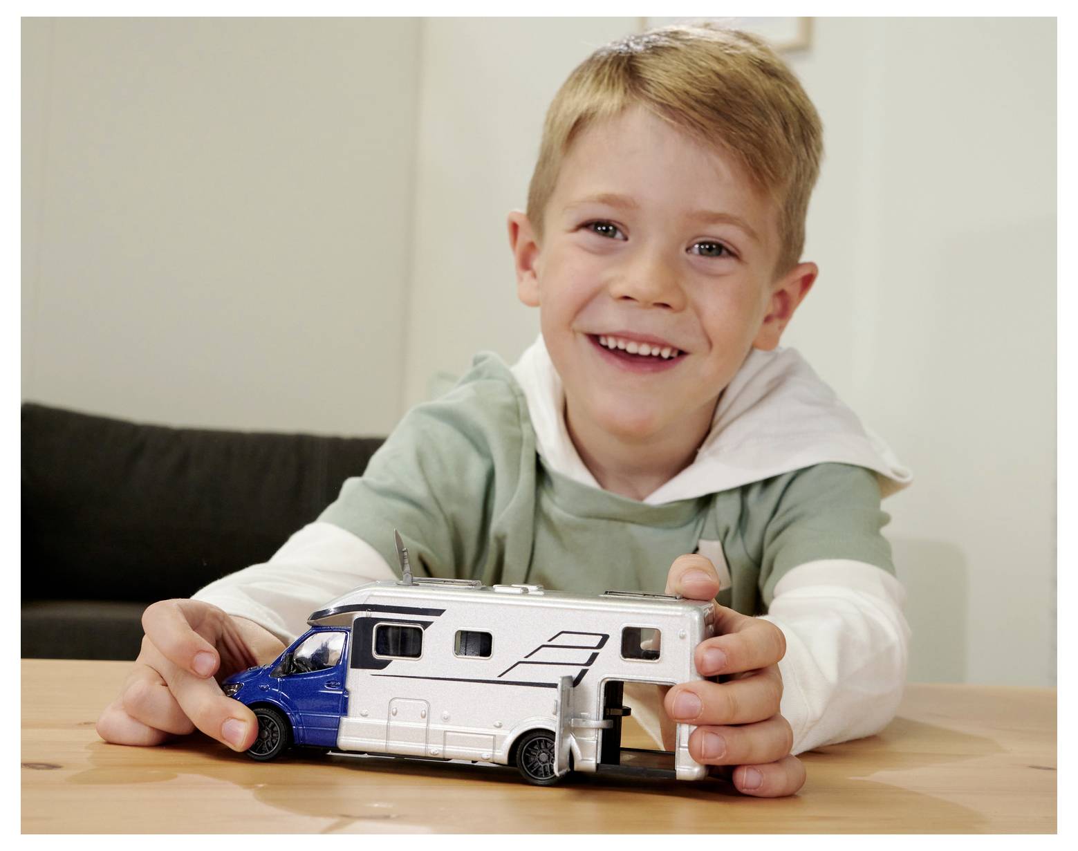 Dziecko uśmiecha się, bawiąc małą zabawkową furgonetką kempingową na drewnianym stole, wyrażając radość i zaangażowanie w zabawę wyobrażeniową.