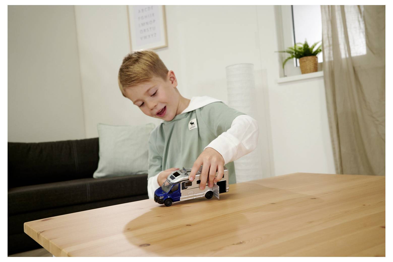 Młody chłopiec radośnie bawi się zabawkowym samochodem na drewnianym stole w przytulnym salonie z sofą i małą doniczkową rośliną przy oknie.