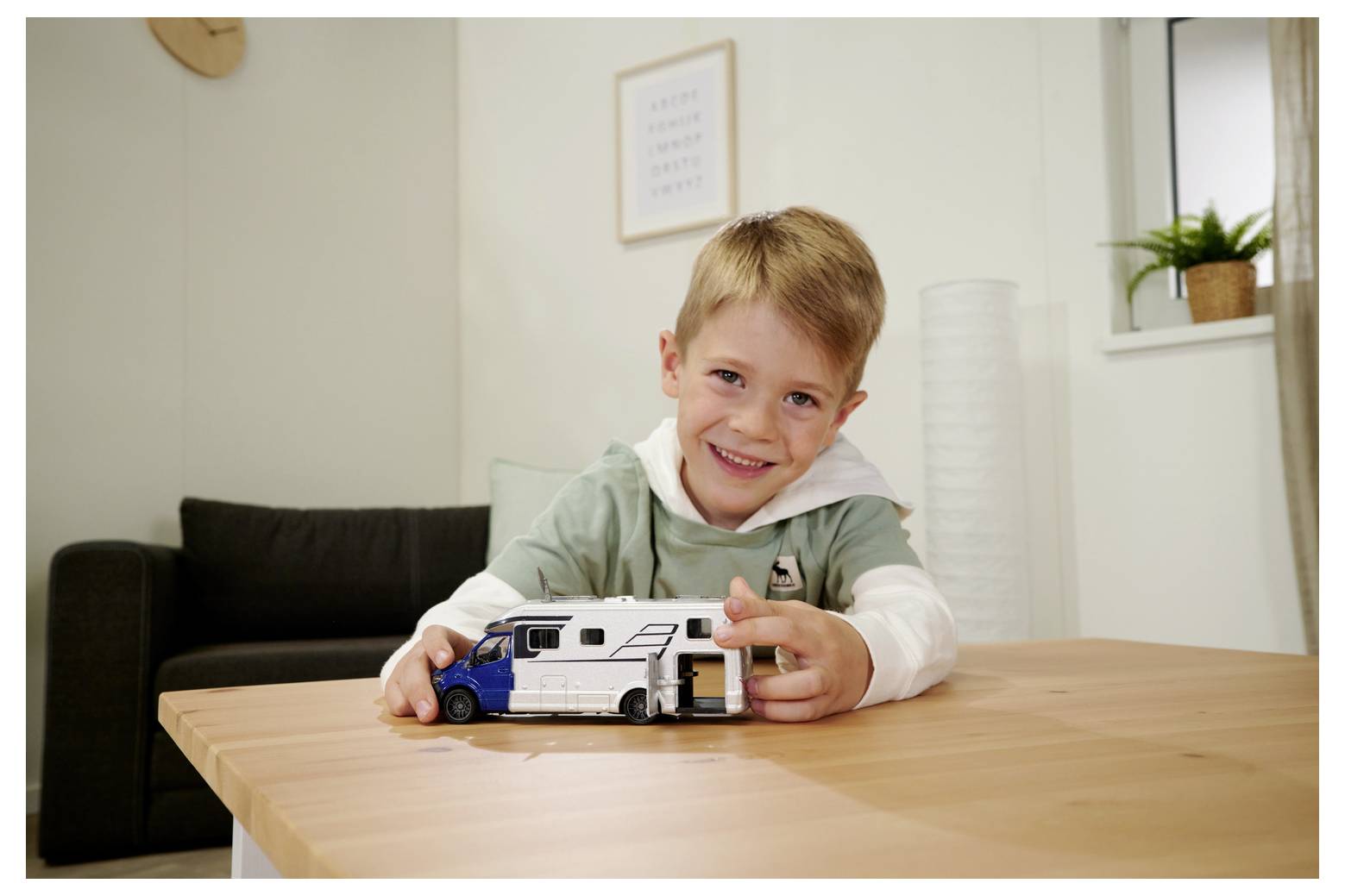 Uśmiechnięte dziecko w zielonej koszulce bawi się zabawkowym kamperem na drewnianym stole we wnętrzu nowoczesnego salonu.