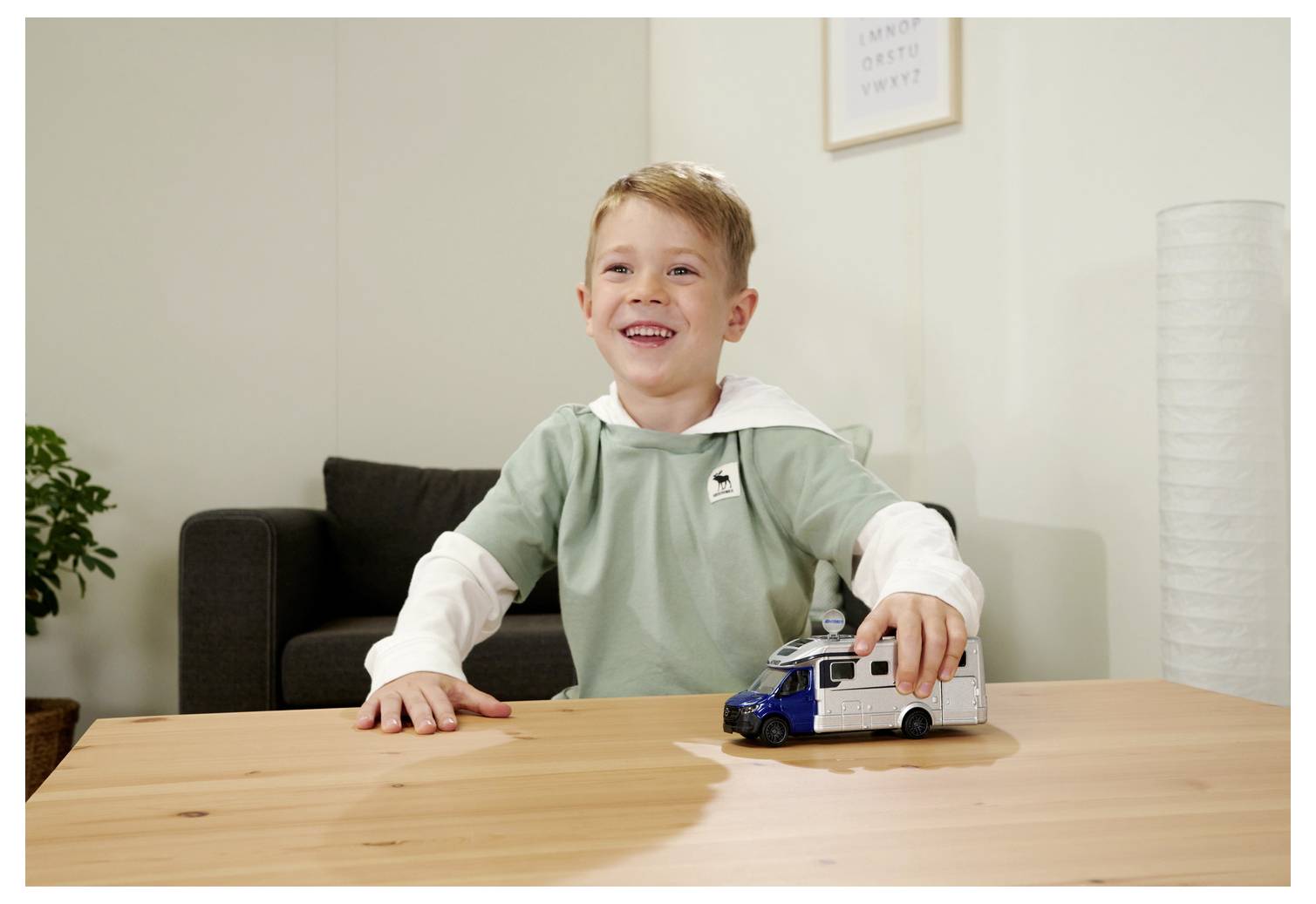 Młody chłopiec uśmiecha się, siedząc przy stole i bawiąc się zabawkowym samochodem dostawczym. W tle widać sofę i oprawiony obraz.