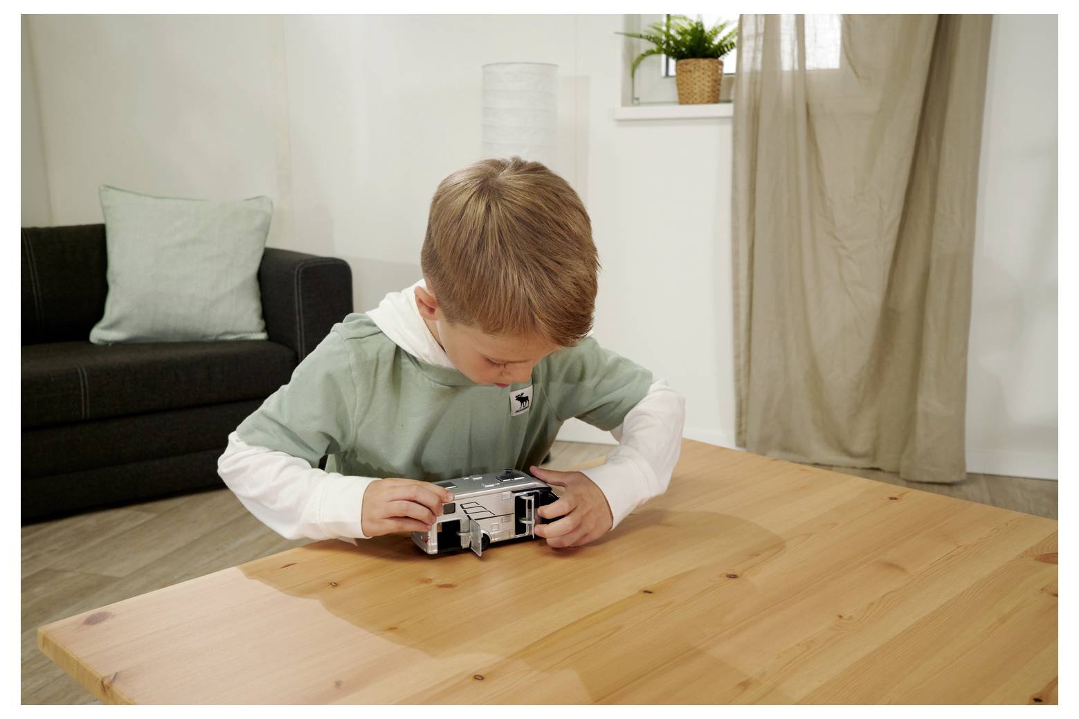 Dziecko bawi się małą zabawkową autobusową na drewnianym stole w salonie, w tle widać sofę, roślinę i zasłonę.