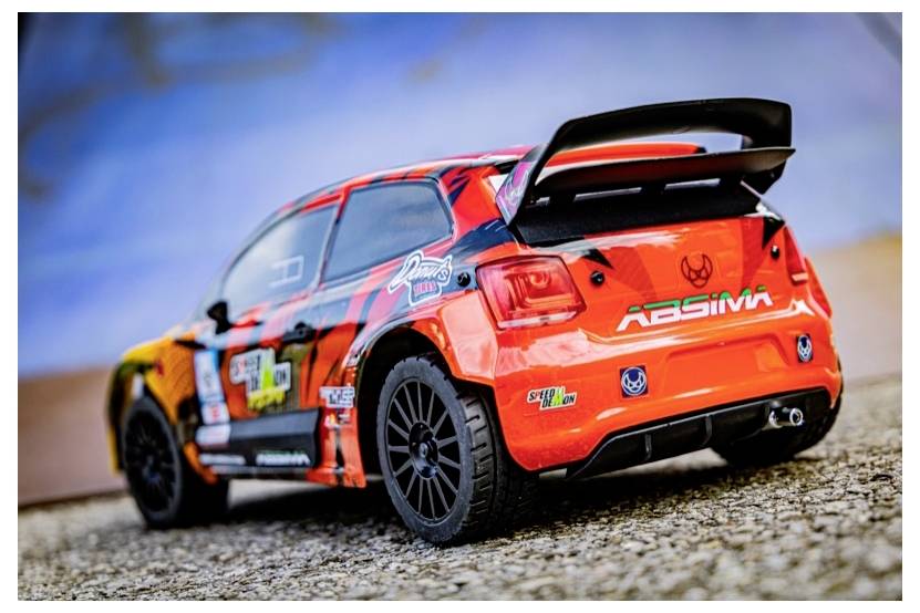 Żywy zabawkowy samochód wyścigowy z pomarańczowymi, czarnymi i czerwonymi wzorami, zaparkowany na teksturowanej powierzchni, prezentujący szczegółowe naklejki i tylny spojler.