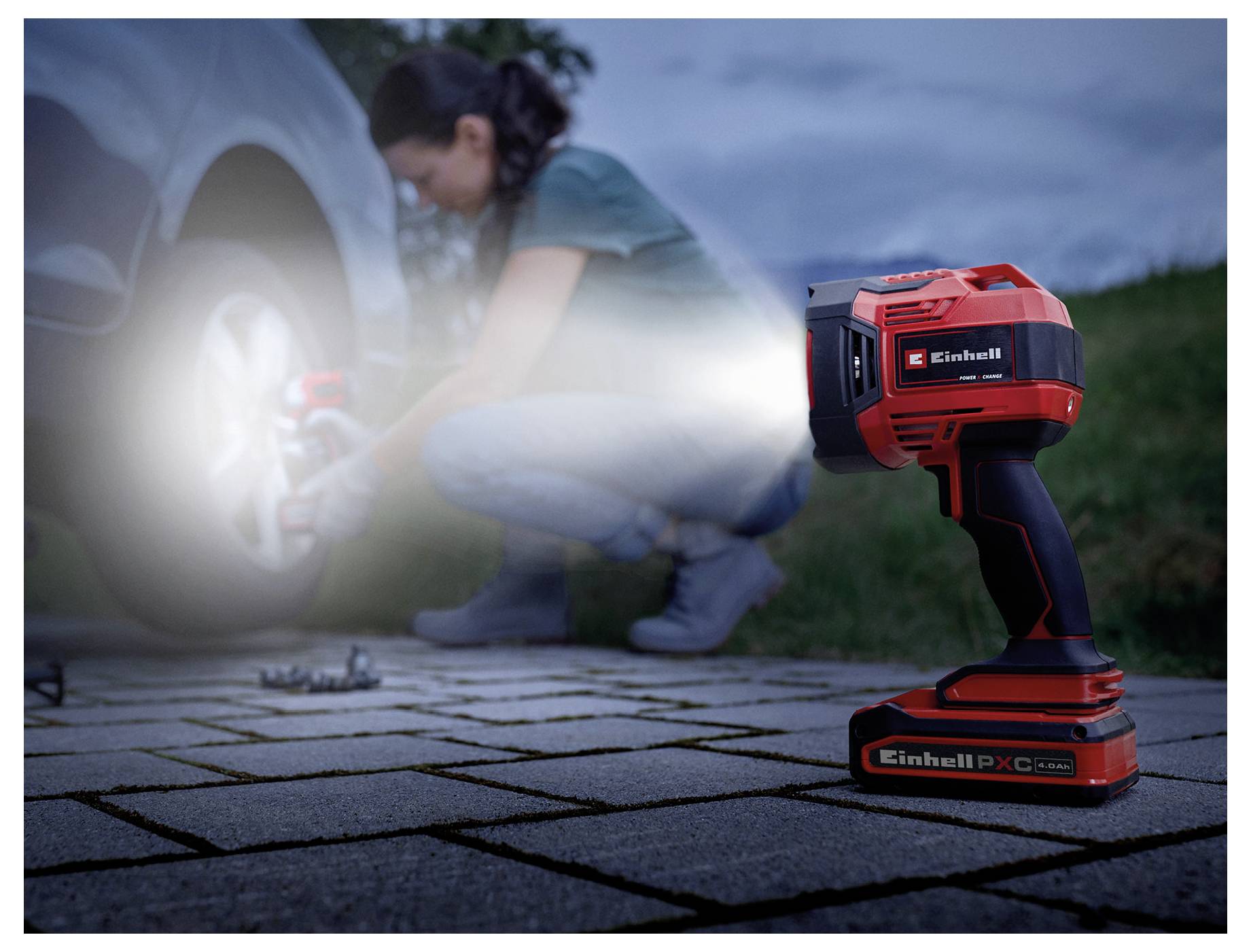 Osoba używająca przenośnego narzędzia elektrycznego do dokręcania koła samochodowego na podjeździe, z naciskiem na emitujące światło narzędzie, podkreślające jego zastosowanie.