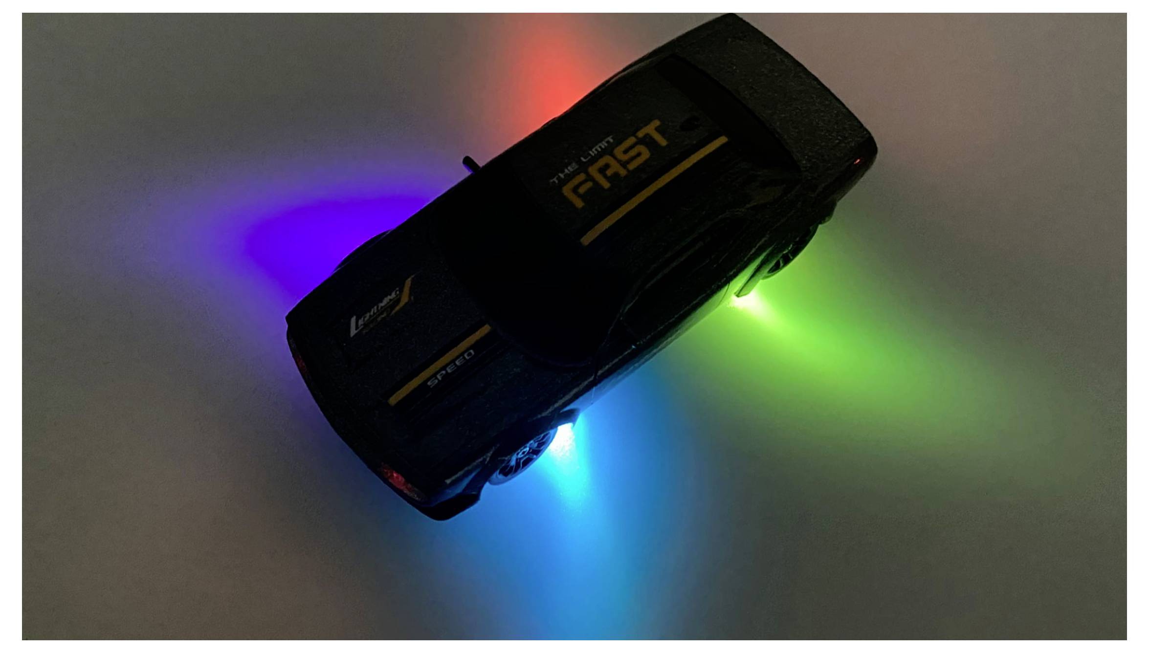 Zabawkowy samochodzik z napisem 'fast' na wierzchu jest podświetlony od spodu czerwonymi, niebieskimi i zielonymi światłami, tworząc kolorową poświatę na ciemnym tle.