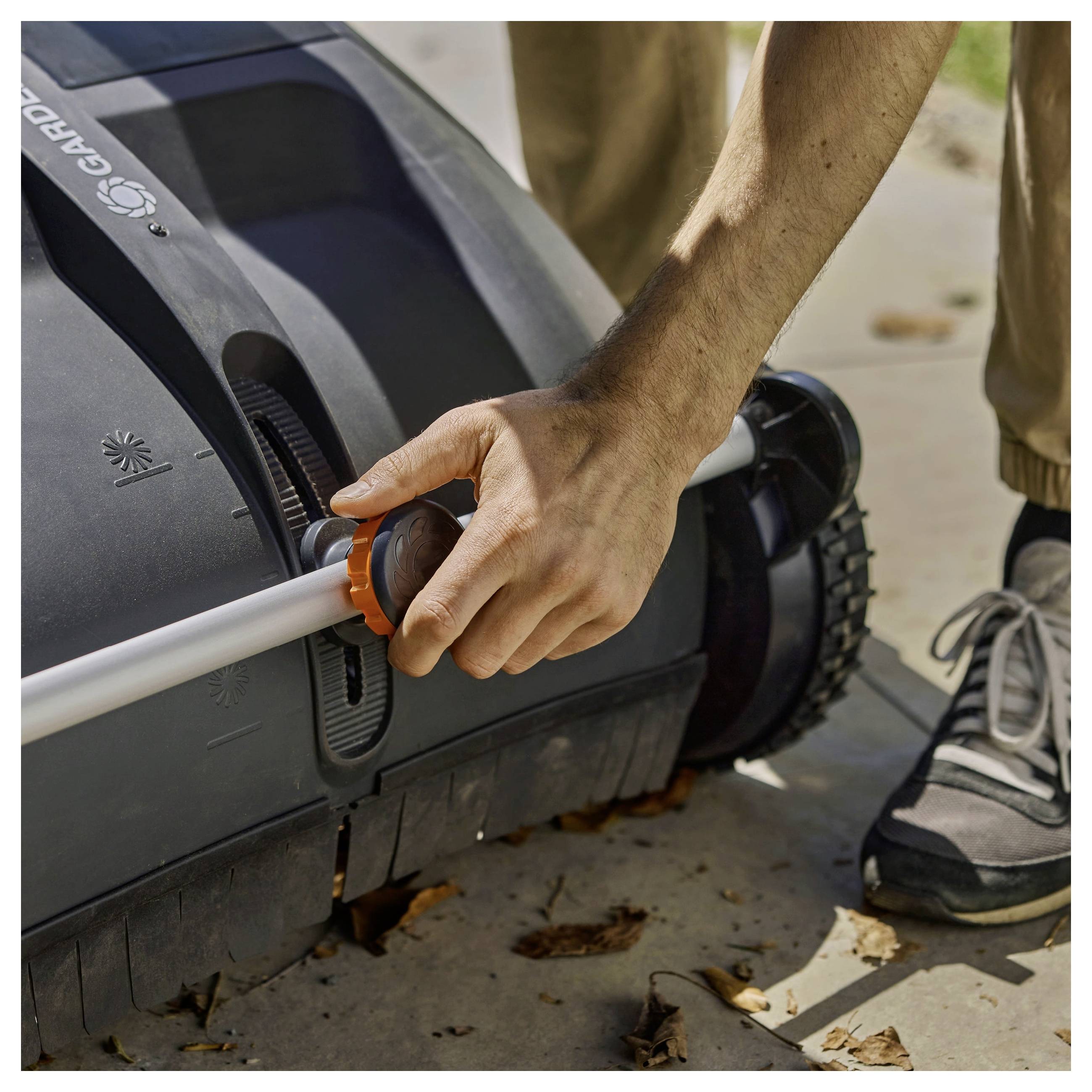 Osoba regulująca wysokość kosiarki do trawy za pomocą pokrętła, stojąca na chodniku z liśćmi rozrzuconymi wokół.